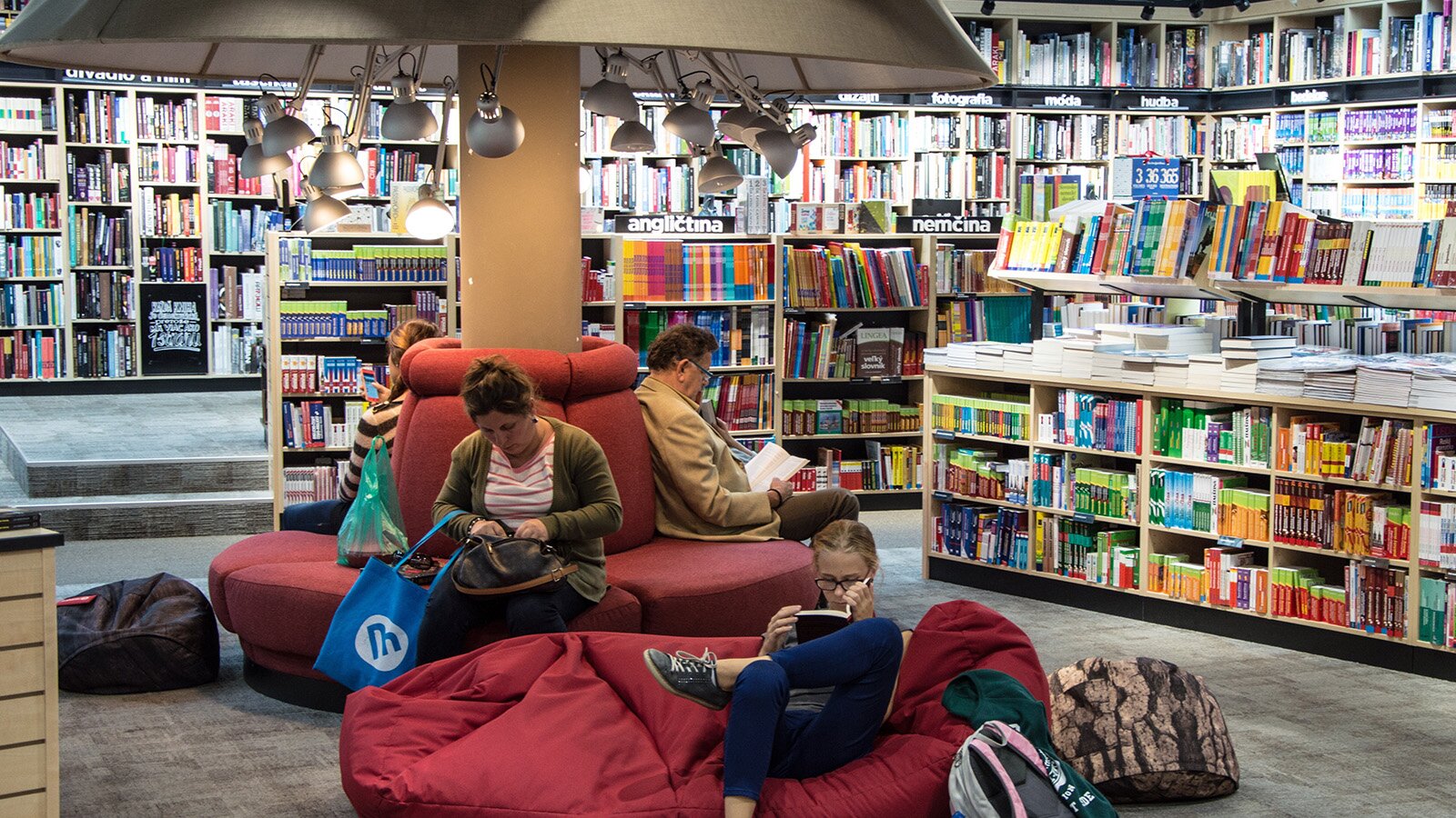 Zdjęcie przedstawia wnętrze księgarni. W centralnej części widoczne jest okrągłe siedzisko usytuowane dookoła kolumny. Siedzisko ma miękkie, czerwone obicia, a nad nim widoczny jest duży abażur, z przymocowanymi od spodu kilkunastoma święcącymi się lampkami. Na siedzisku siedzą trzy osoby, które czytają książki. Przed okrągłym siedziskiem widoczny jest duży czerwony puf stojący na podłodze, na którym siedzi dziewczyna i czyta książkę. W tle widoczne są niskie i wysokie regały wypełnione książkami. Na niskich regałach książki są poustawiane na półkach i położone na górnych powierzchniach regałów.