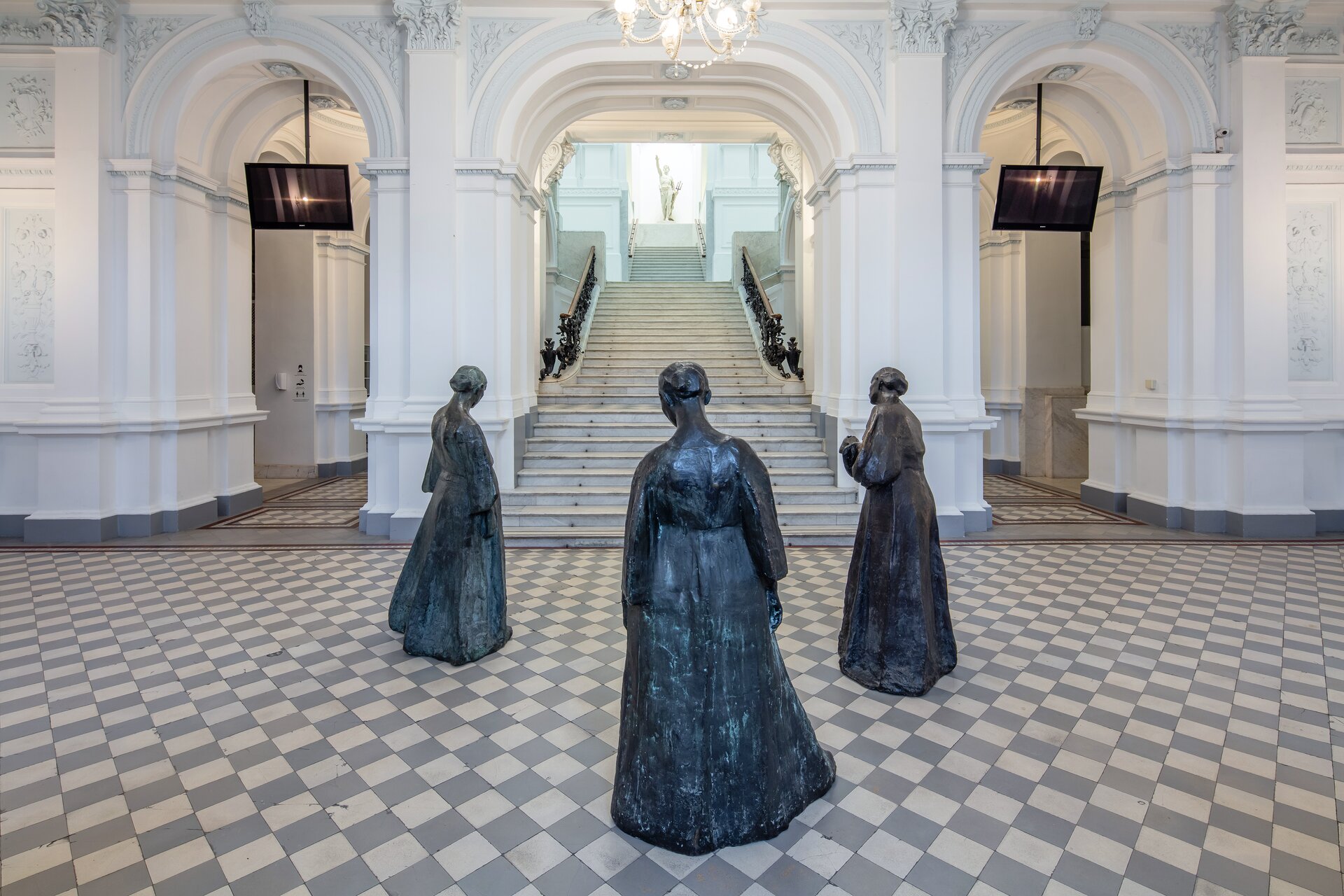 Fotografia przedstawia pomieszczenie w galerii sztuki, w którym na środku znajdują się trzy czarne rzeźby, ukazujące kobiety. Posągi odwrócone są tyłem do obserwującego. Jedna z kobiet stoi bliżej, dwie pozostałe tworzą drugi plan fotografii. Dalej znajdują się szare schody, prowadzące na piętro, gdzie znajduje się posąg. Rzeźba ma uniesioną prawą rękę w górze. Na białych ścianach ukazane są ozdobne ornamenty.