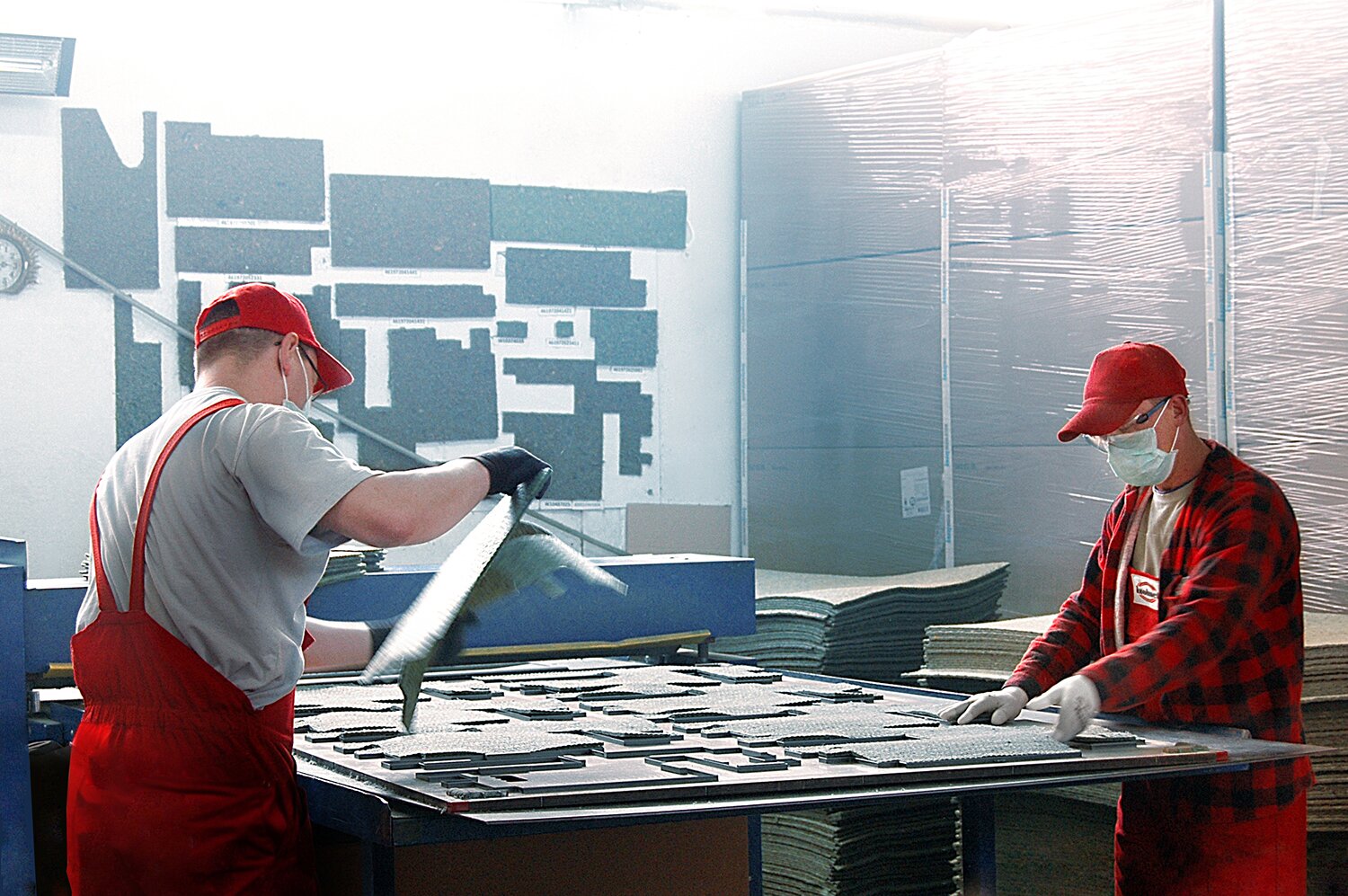 Zdjęcie przedstawia dwóch pracowników w strojach roboczych, maskach na twarzy i okularach ochronnych. Sprawdzają oni elementy izolacji na dużym stole.