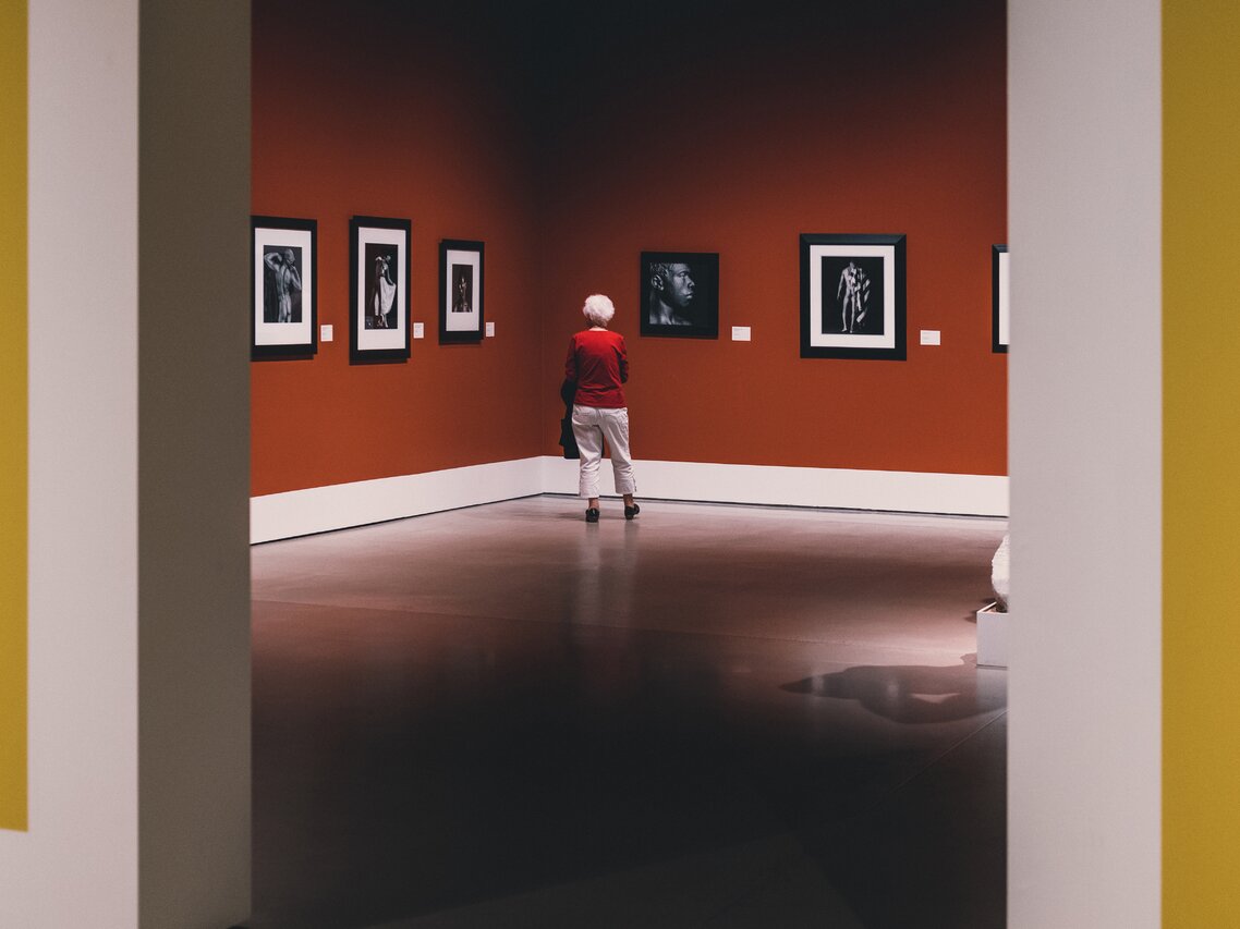 Fotografia barwna. Sala galerii pomalowana na rdzawoczerwony kolor. Na ścianach wiszą czarno-białe zdjęcia przedstawiające czarnoskórych mężczyzn. Przed jednym z nich stoi tyłem do nas siwa, krótkowłosa kobieta. Ubrana w czerwony sweter, białe spodnie i czarne buty.