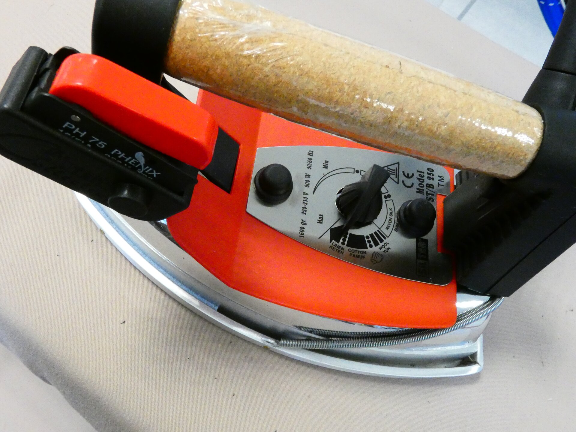 Zdjęcie przedstawia metalowe żelazko z pomarańczowymi elementami i drewnianą rączką. Na nim znajdują się pokrętła.