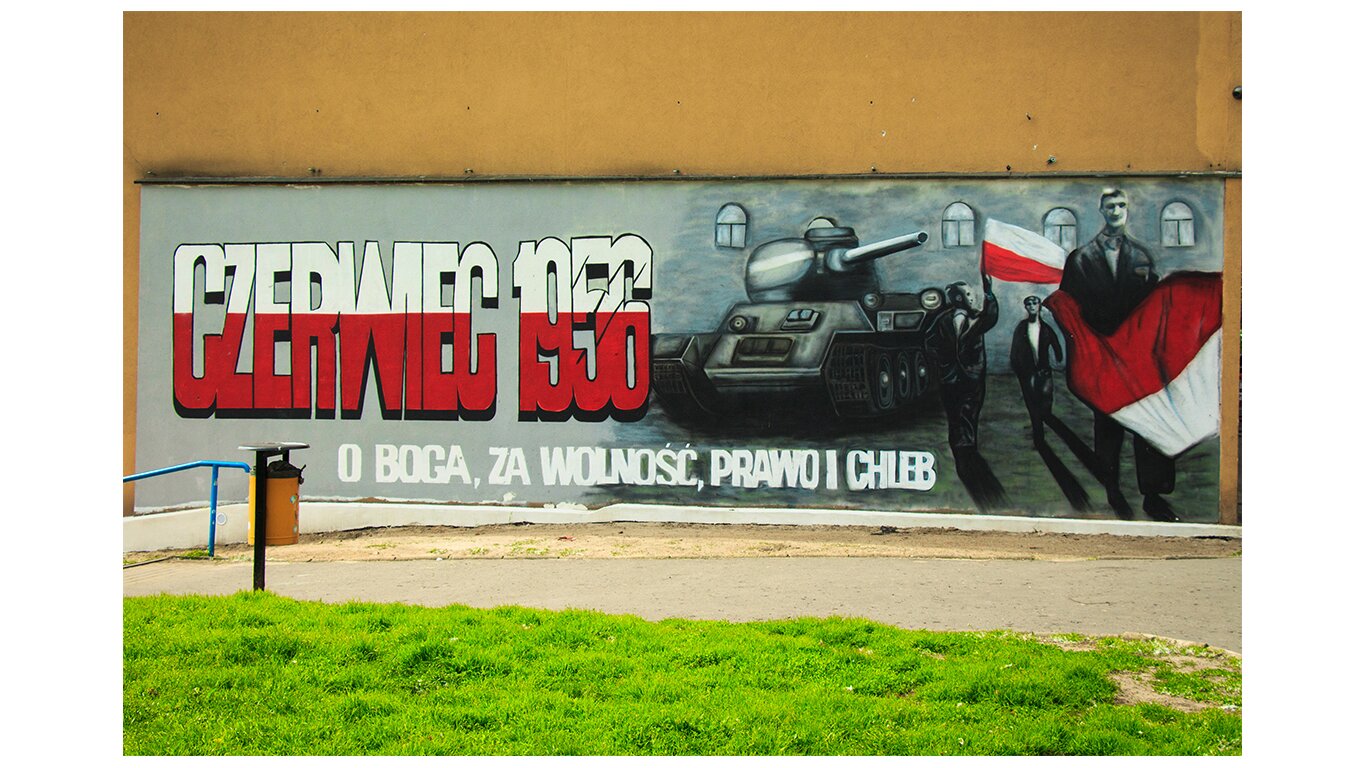Fotografia barwna przedstawiająca mural upamiętniający wydarzenia z czerwca 1956 r. w Poznaniu. Po lewej biało-czerwony napis Czerwiec 1956, po prawej czołg skierowany lufą w stronę robotników niosących biało-czerwone flagi. Na dole napis: O Boga, za wolność, prawo i chleb.