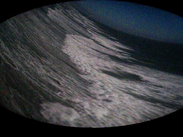 Zdjęcie wykonane kamerą otworkową, czyli camerą obscura. Przedstawia fale morskie sfotografowane przez luk statku. Zdjęcie jest zakrzywione, nieostre na brzegach. 