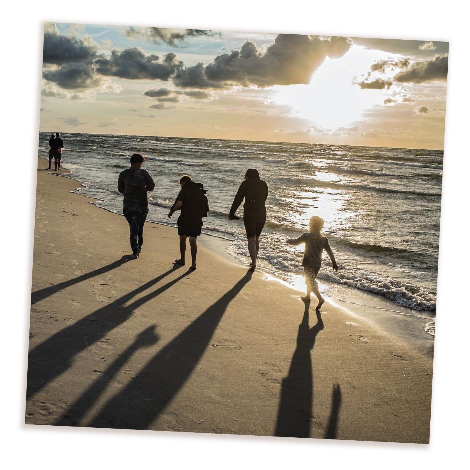 Zdjęcie przedstawia grupę ludzi idących po plaży przy brzegu  podczas zachodu słońca. Jeden mężczyzna stoi na brzegu mocząc stopy w morzu. 