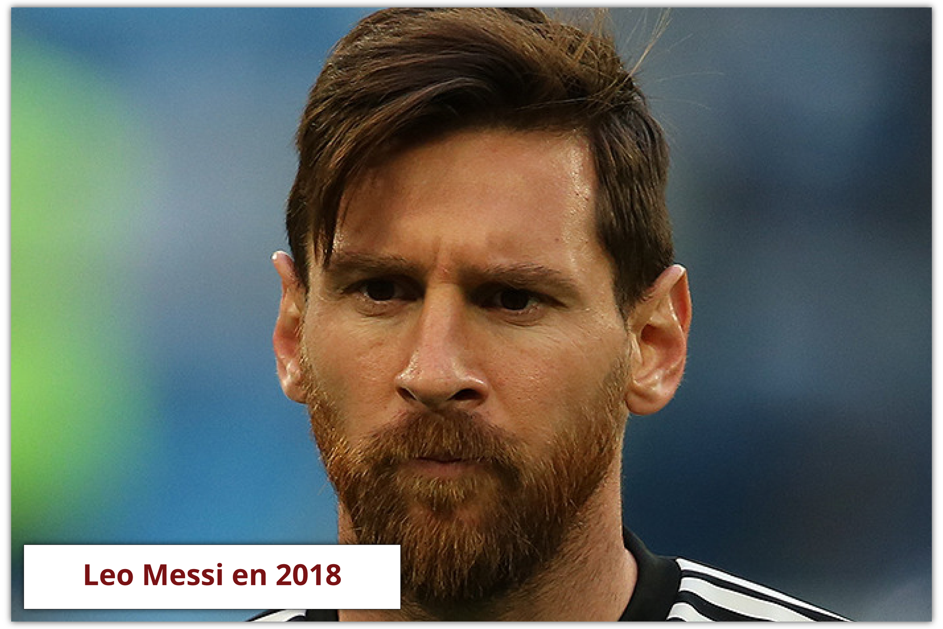 Na zdjęciu widoczny jest mężczyzna z brodą i brązowymi włosami. Ma na sobie sportową koszulkę. W tle widoczne jest rozmazane, zielono‑niebieskie tło. Mężczyzna ma poważny wyraz twarzy. Podpis zdjęcia: Leo Messi en 2018.