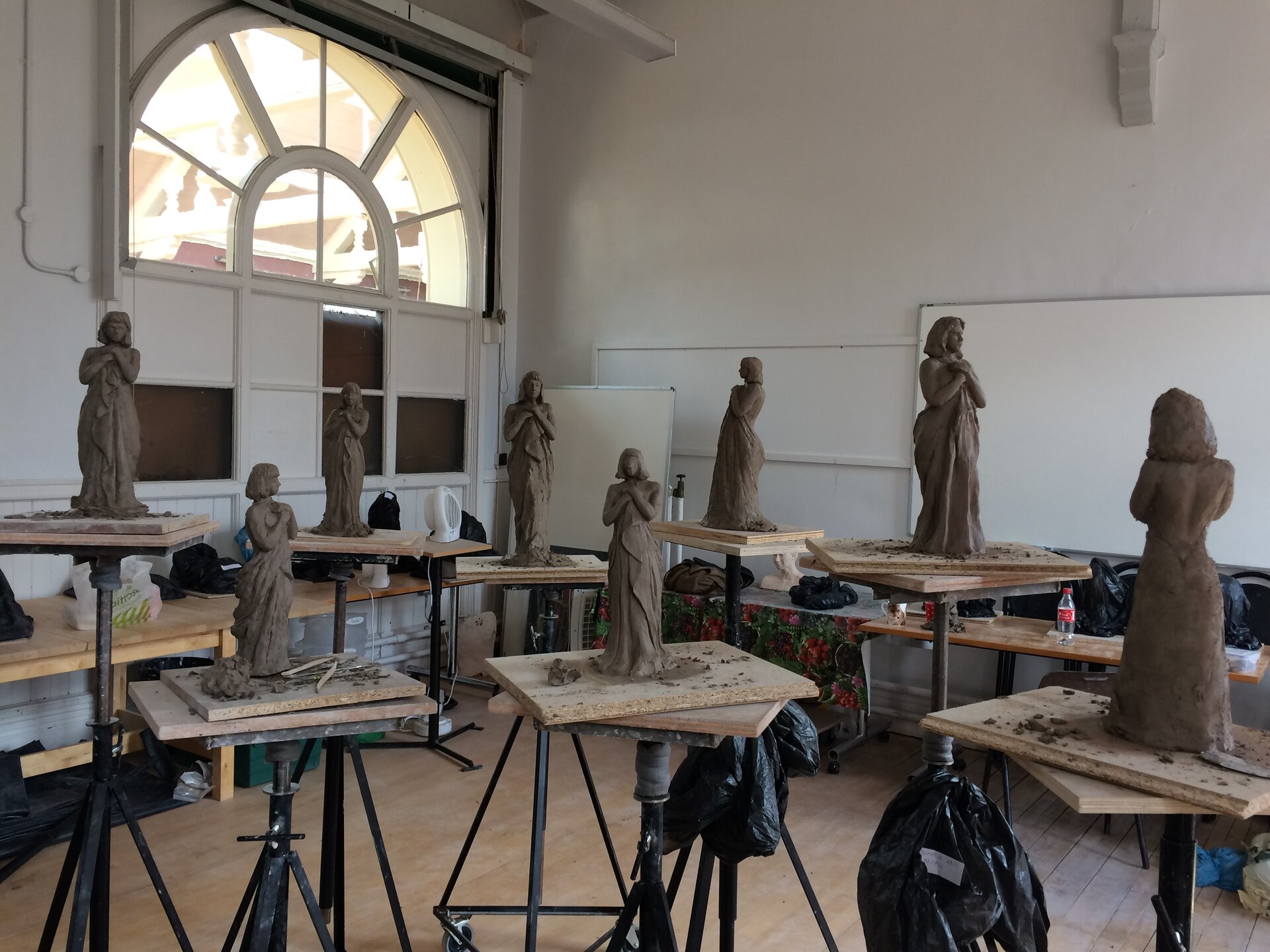 Fotografia przedstawia pracownię artystyczną, w której znajdują się rzeźby wykonane z gliny. W dużym pomieszczeniu widocznych jest osiem stojaków, na każdym z nich stoi rzeźba. Rzeźby przedstawiają krótkowłosą kobietę w długich szatach, krzyżującą przed sobą dłonie. W lewym górnym rogu fotografii widoczne jest okno, przez które do pomieszczenia wpada światło. Stojaki mają czarne podstawy oraz jasne, drewniane, prostokątne blaty. W tle widoczne są dwie białe tablice na tle białych ścian. 