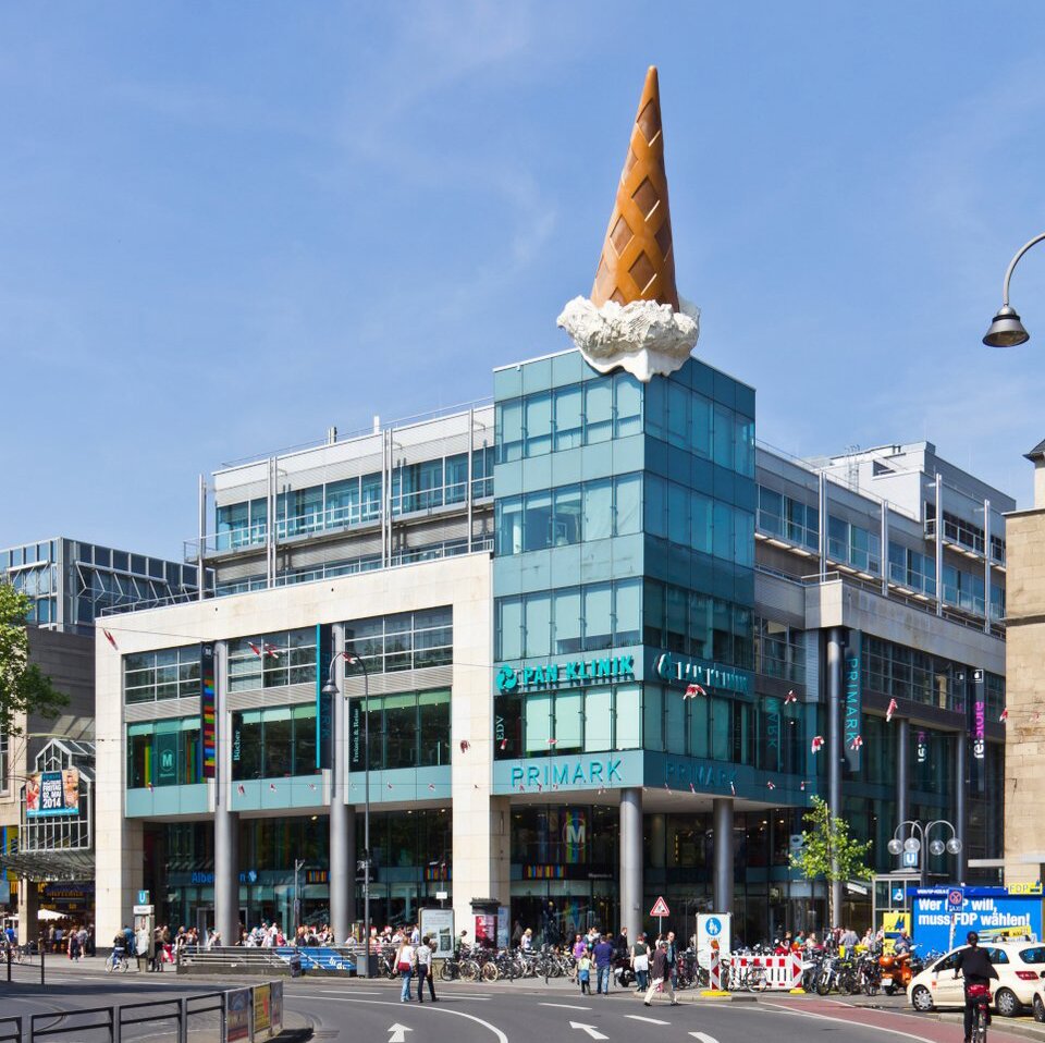 Zdjęcie przedstawia budynek centrum handlowego przy ruchliwej ulicy, na którego dachu umieszczono rzeźbę odwróconego loda w rożku.