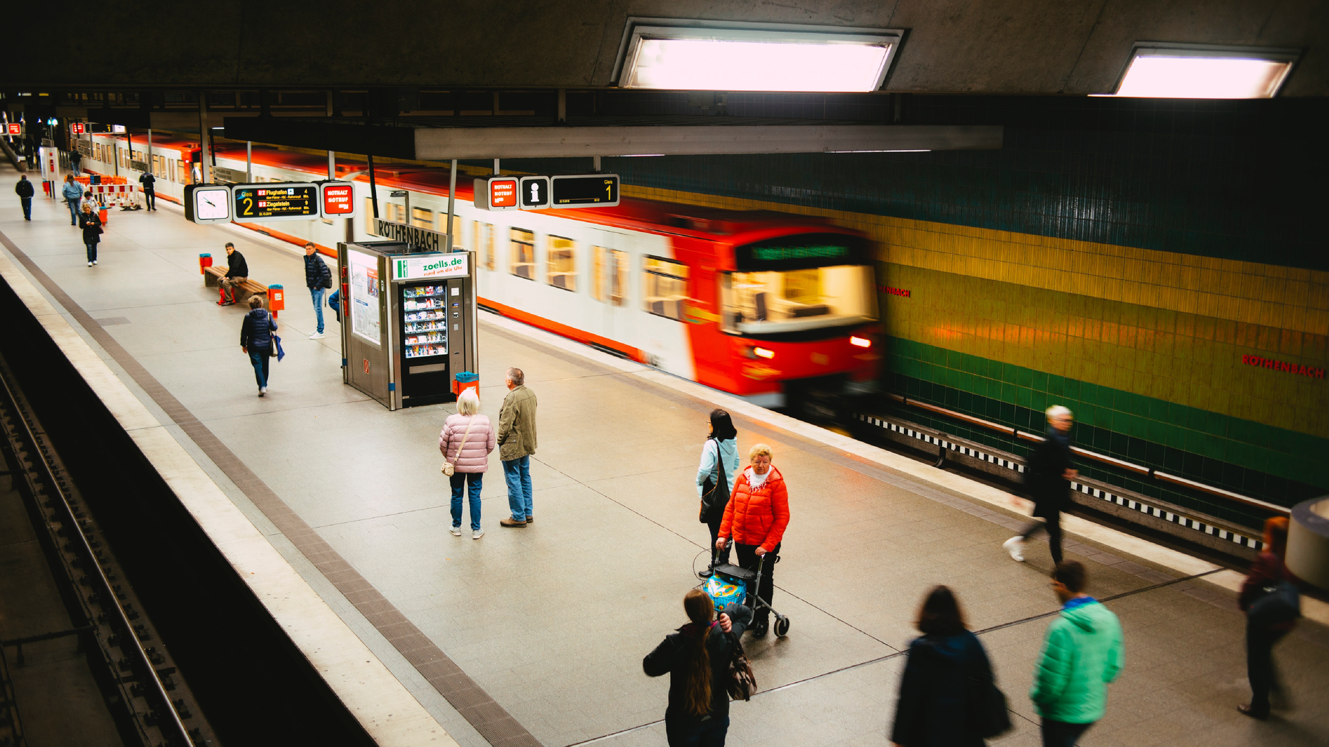 Zdjęcie zrobione z góry przedstawia peron na stacji metra. Na peronie znajdują się ludzie, widoczny jest automat samoobsługowy z przekąskami. Na prawym torze znajduje się pociąg, lewy tor jest pusty. Nad peronem widoczne są tablice z rozkładem jazdy oraz zegar.