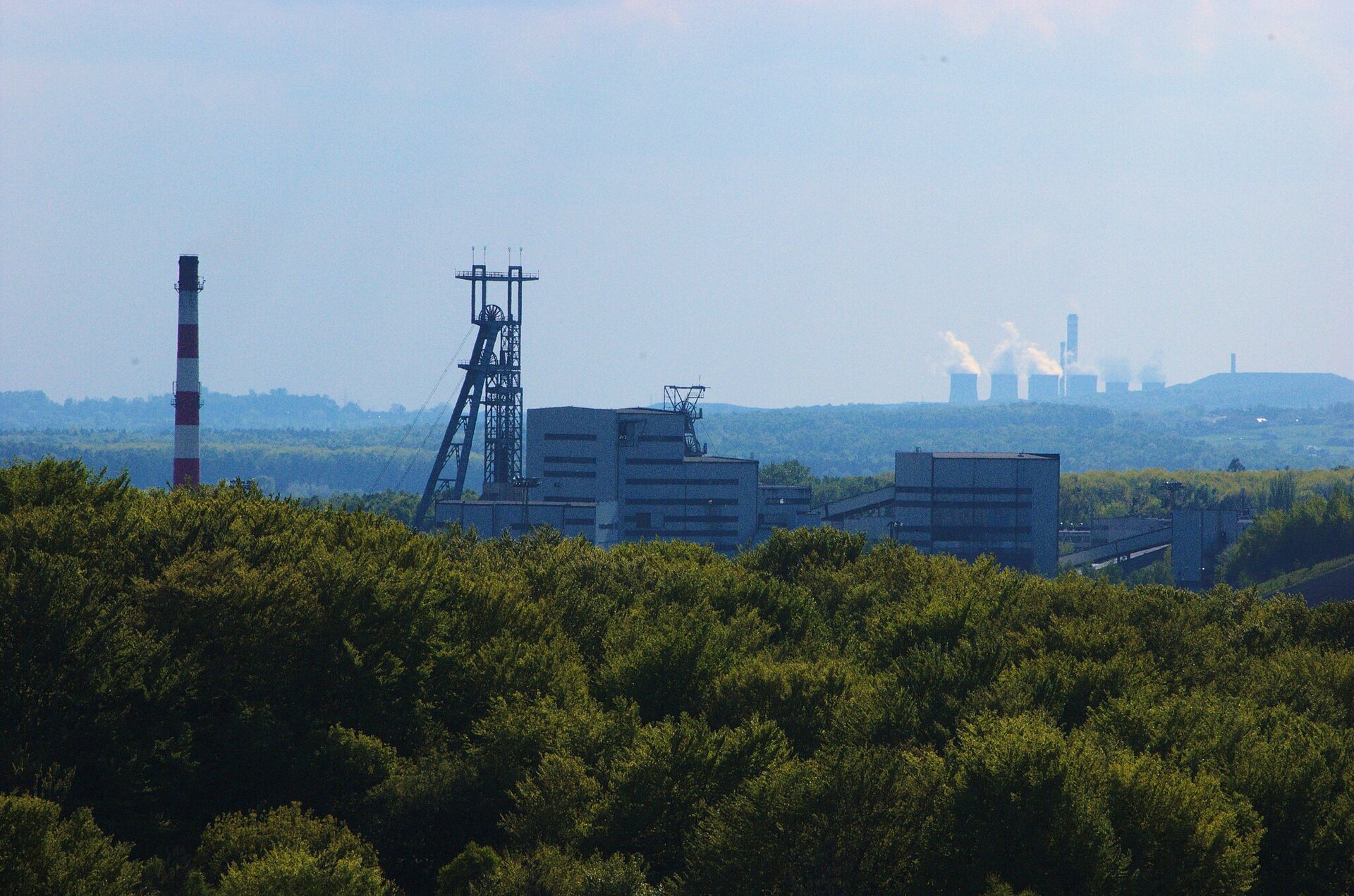 Fotografia prezentująca górujące nad otaczającym je lasem zabudowania kopalni. Kilkupiętrowe budynki, obok nich wysoki metalowy szyb oraz wysoki komin. Na drugim planie szerokie i duże dymiące na biało kominy elektrowni. Dookoła las.