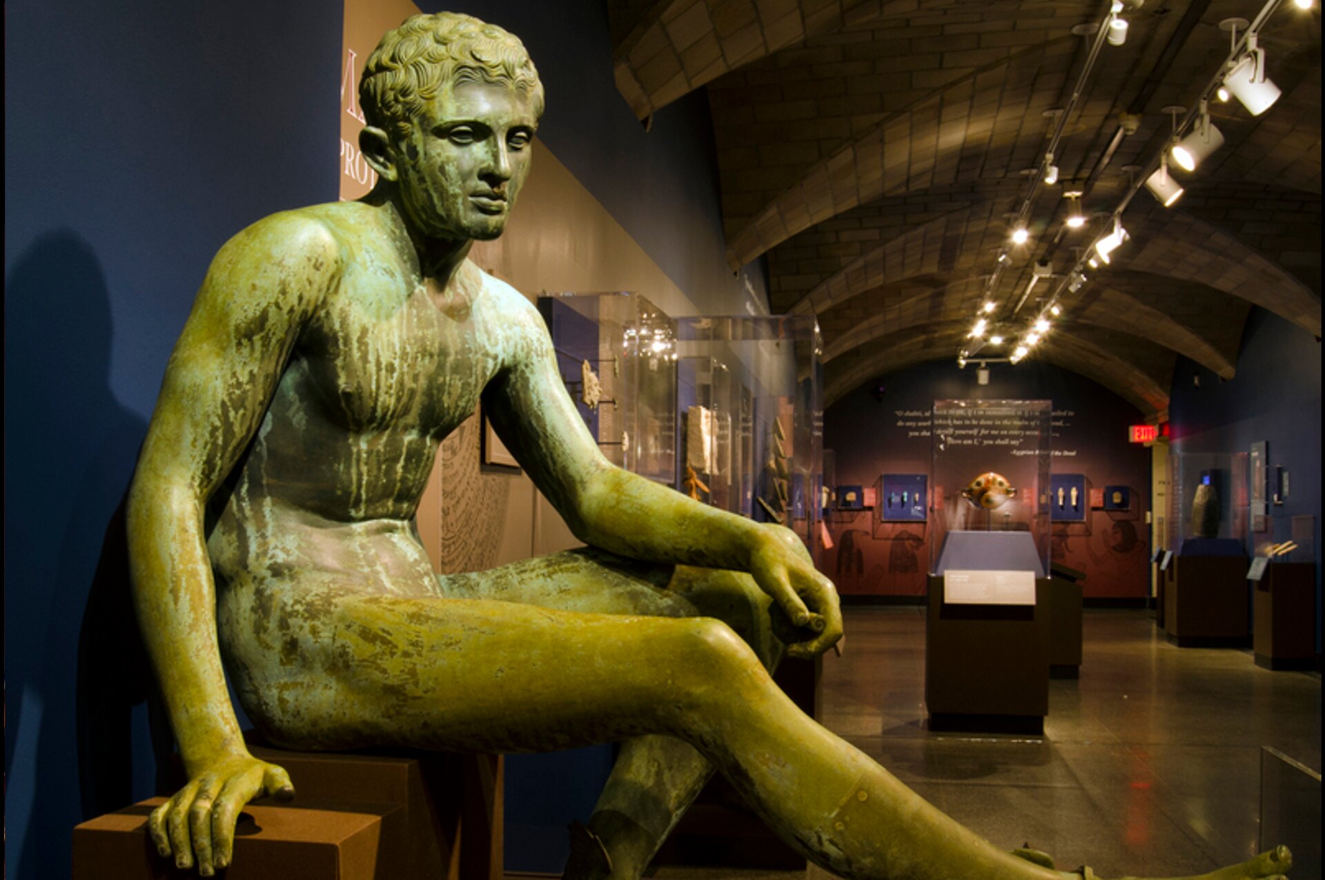 Zdjęcie przedstawia rzeźbę nagiego, młodego mężczyzny. Mężczyzna siedzi na postumencie. Rzeźba znajduje się w muzeum, w tle są eksponaty w szklanych gablotach.