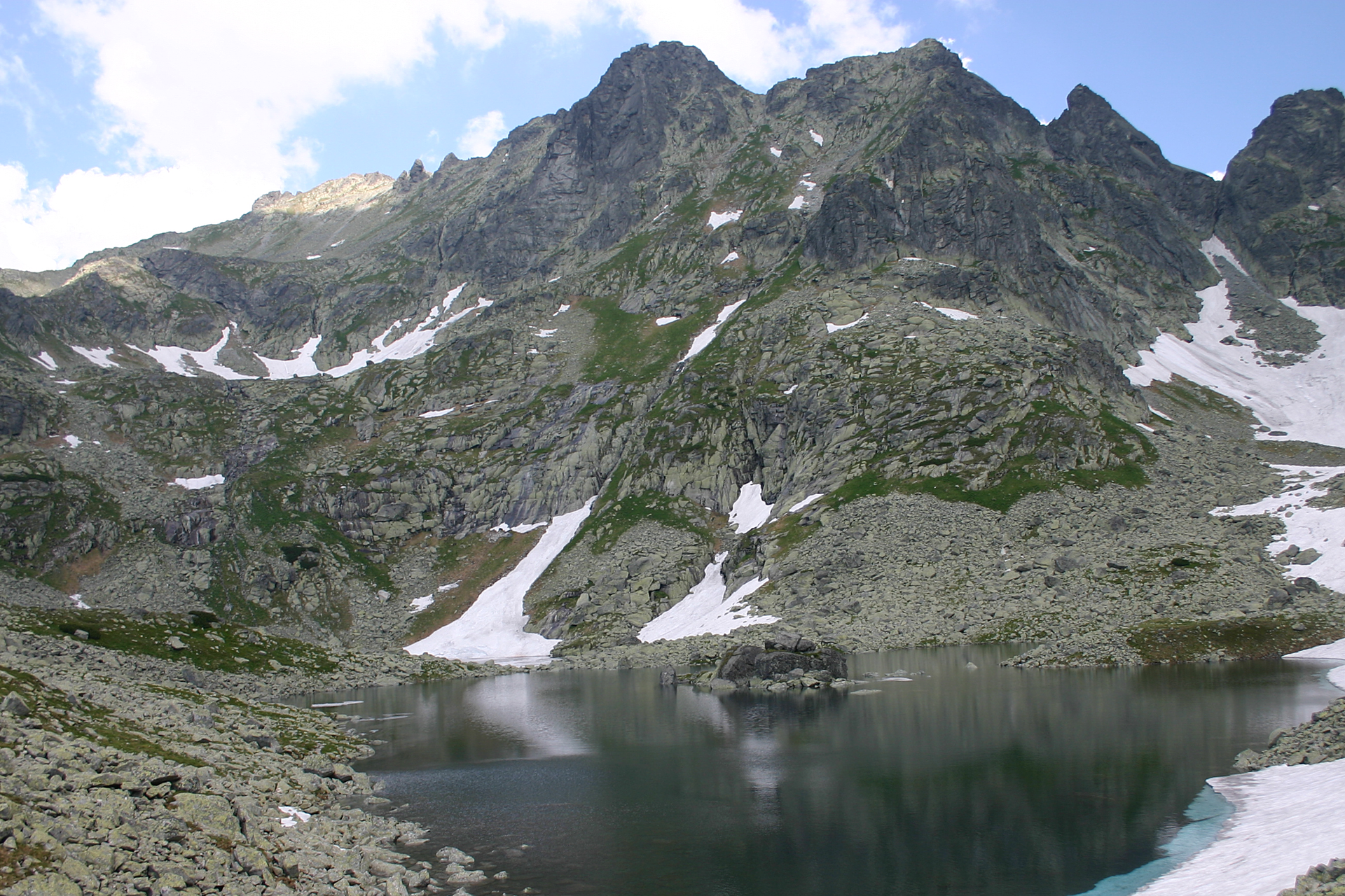 Fotografia ukazująca panoramę gór wysokich nad jeziorem, Morskim Okiem. Widoczne cztery szczyty górskie, z których najwyższy po lewej stronie, to Rysy. Stoki szczytów gdzieniegdzie są pokryte śniegiem.