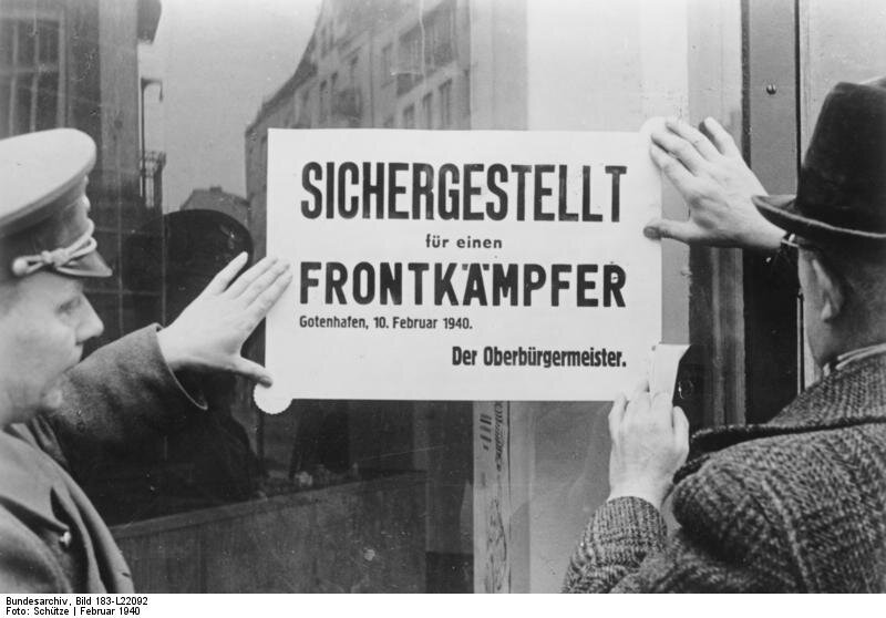 Zdjęcie przedstawia dwóch mężczyzn mocujących dużą kartkę na witrynie sklepowej. Jeden z nich jest w niemieckim mundurze, drugi w ubraniu cywilnym. Na kartce widnieje napis w języku niemieckim: Sicherestellt für einen Frontkämpfer. Gotenhafen, 10. Februar 1940. Der Oberbürgermeister.