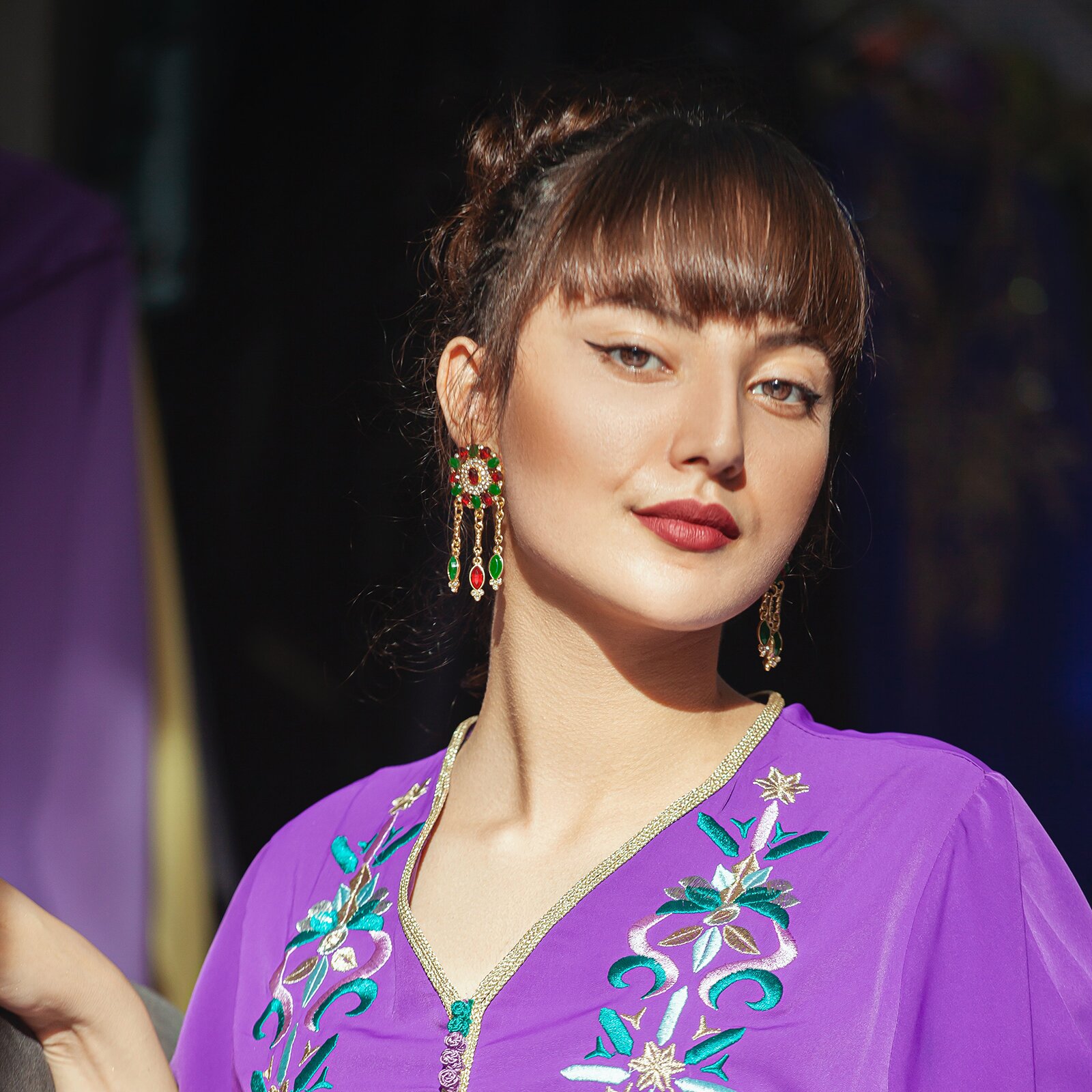 Zdjęcie przedstawia młodą kobietę o brązowych, upiętych włosach z grzywką. Ma brązowe, duże oczy. Ma również pomalowane usta. Nosi wyszywaną bluzkę i kolczyki.