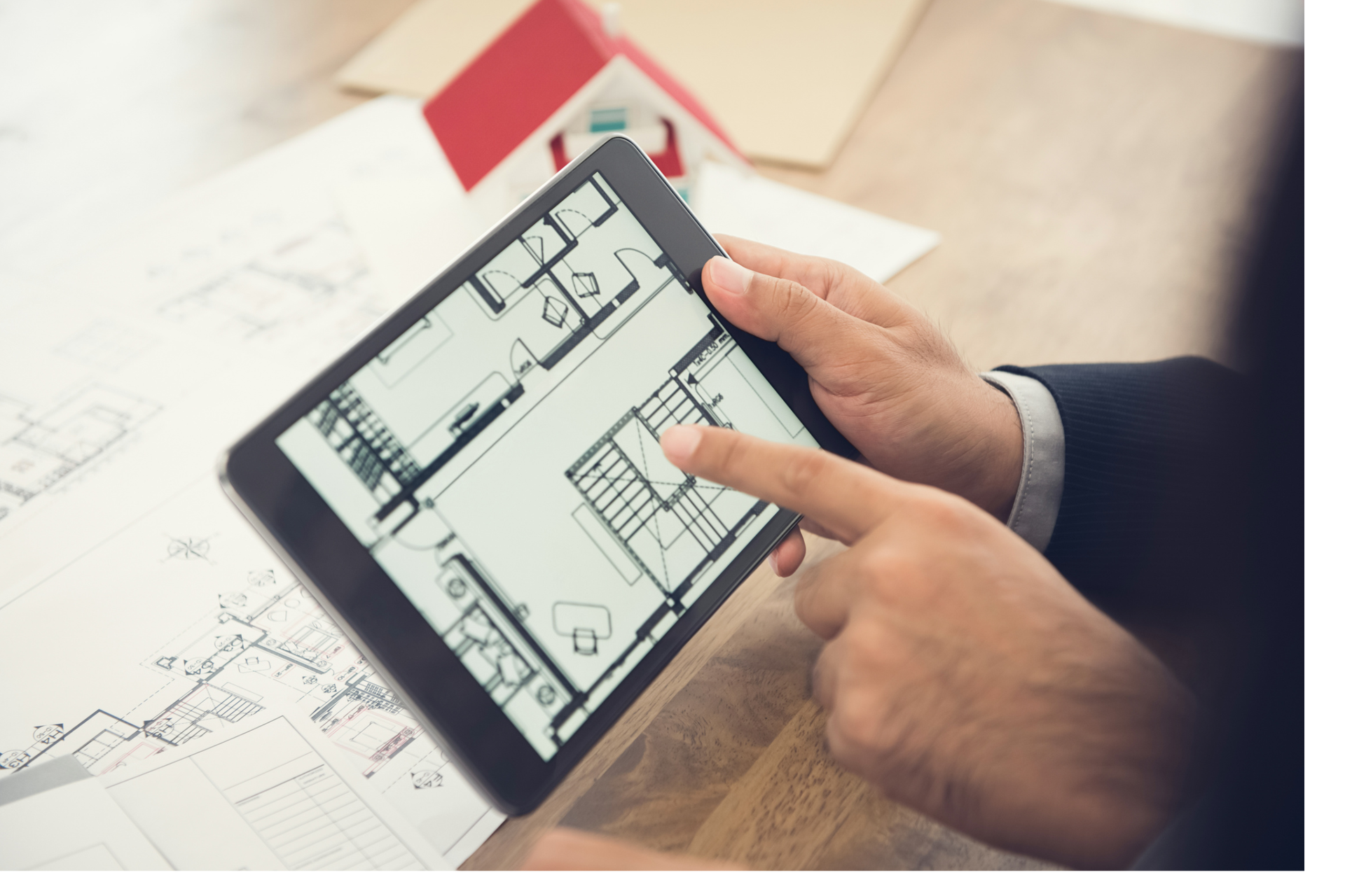 Zdjęcie przedstawia tablet trzymany przez męską dłoń w garniturze, na którym wyświetlany jest plan budynku. Druga ręka mężczyzny wskazuje na ekran. W głębi na stole leżą plany architektoniczne wydrukowane na papierze. 