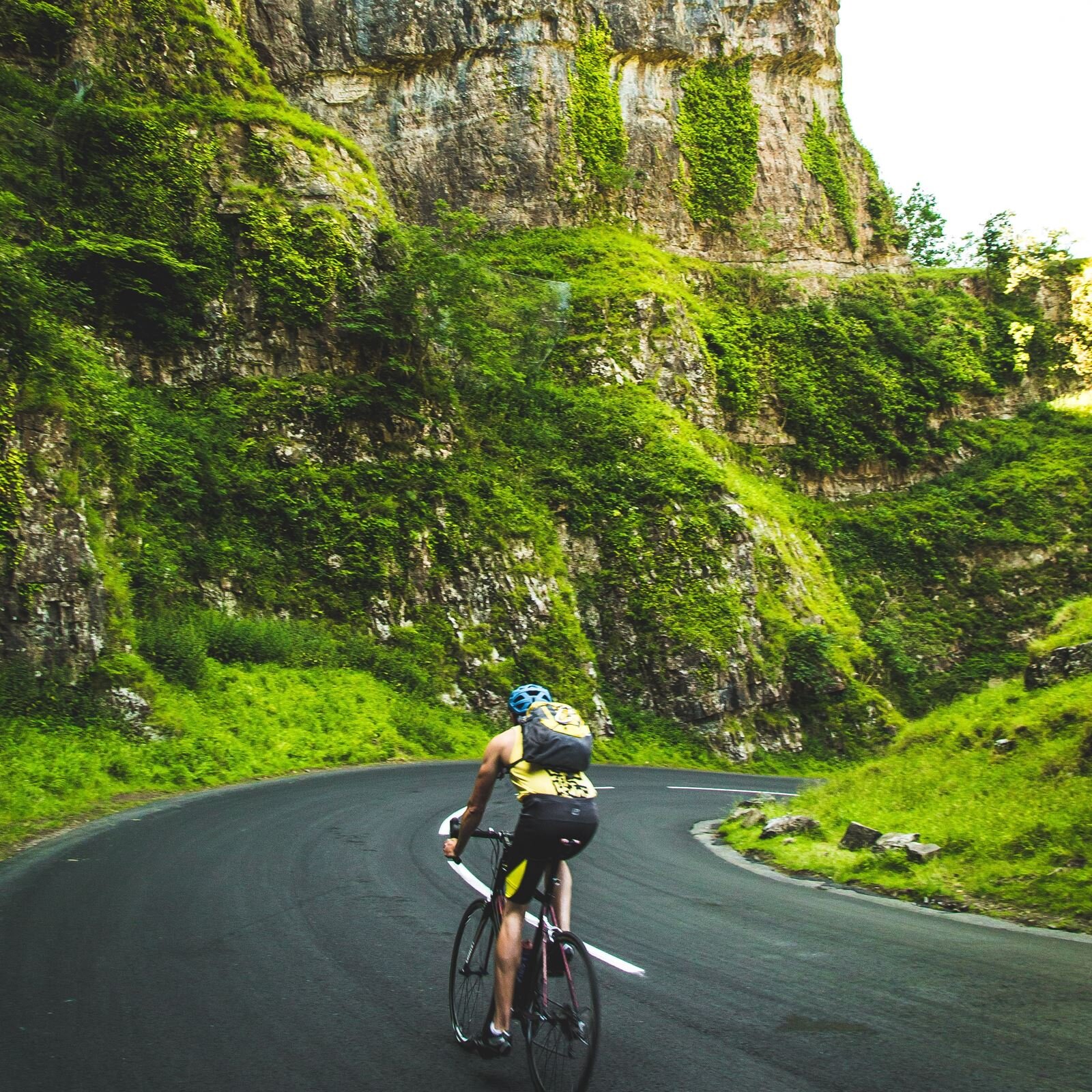Zdjęcie przedstawia mężczyznę, w ujęciu od tyłu, jadącego na rowerze po krętej asfaltowej drodze. Mężczyzna ma na sobie strój kolarski: krótkie, czarne leginsy, żółtą koszulkę na ramiączkach, niebieski kask ochronny. Na plecach ma założony żółto‑czarny plecak. Porusza się w dół drogą skręcającą w prawo. Po obu stronach drogi widoczne są skaliste góry, które w dolnych i środkowych partiach porośnięte są bujną, intensywnie zieloną roślinnością.