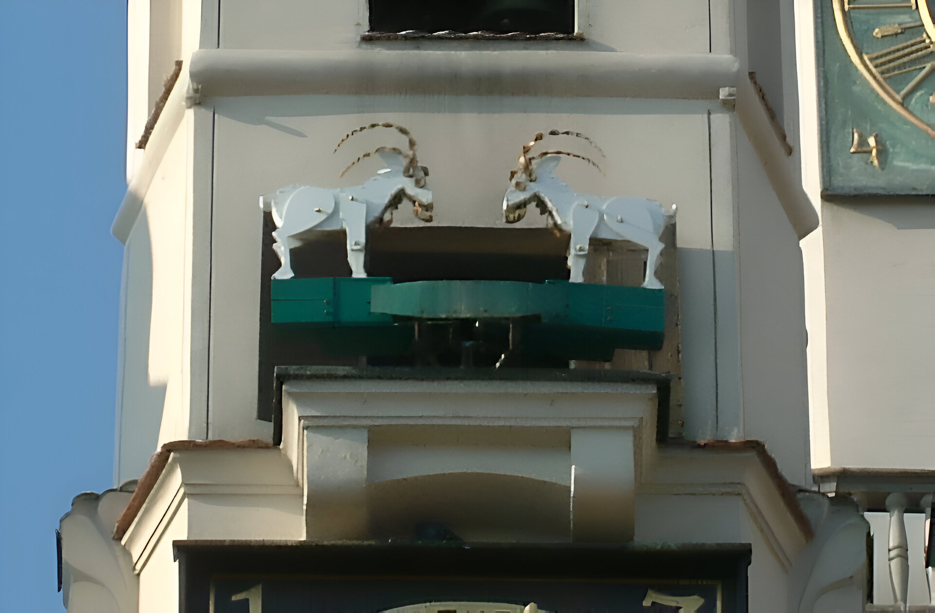 Zdjęcie przedstawia zbliżenie na wieżę ratusza. Na środku znajdują się dwa drewniane koziołki ustawione naprzeciw siebie. W prawym górnym rogu widać fragment zielono‑złotego zegara, a po lewej stronie fragment błękitnego nieba.