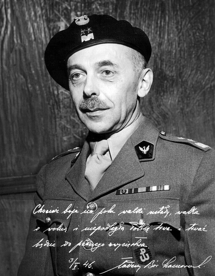 Zdjęcie przedstawia dojrzałego mężczyznę w generalskim mundurze z okresu II wojny światowej. Mężczyzna ma pociągłą twarz, wyrazistą oprawę oczu i prosty nos. Jest ostrzyżony krótko przy skórze, ma wąsy. Na głowie ma beret z orłem i stopniem generała dywizji. Na dole zdjęcia znajduje się odręczny podpis: Chociaż boje na polu walki ustały, walka o wolną i niepodległą Polskę trwa i trwać będzie do pełnego zwycięstwa. 3 maja 1946 Tadeusz Bór Komorowski.