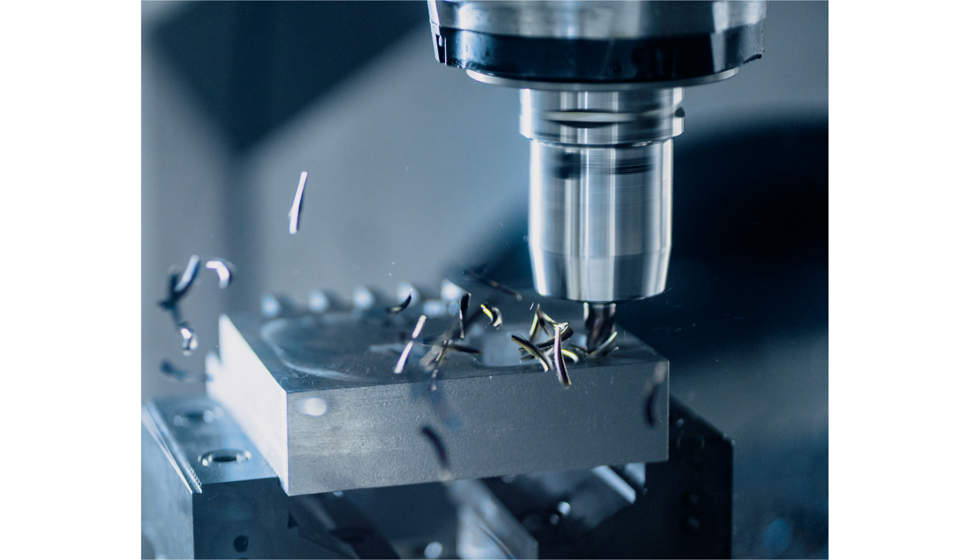 Fotografia przedstawia wiertło w trakcie wiercenia otworu w metalowym bloku, umieszczonym w zacisku. Wiertło osadzone w grubym, pionowym, metalowym walcu. Spod wiertła wylatują metalowe wióry.