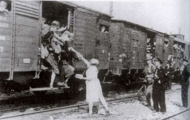 Zdjęcie przedstawia scenę na stacji kolejowej w Pruszkowie, skąd odjeżdża pociąg z wysiedleńcami. Z otwartego wagonu towarowego wychylają się ludzie, aby odebrać z rąk siostry PCK chleb. Kobieta trzyma bochenek razowca, stawiając stopę na wyciągniętej belce wagonowej. Z innych wagonów wychylają się ludzie. Scenie odjazdu przyglądają się kolejarze oraz pracownik niemieckiej administracji Generalnego Gubernatorstwa. 