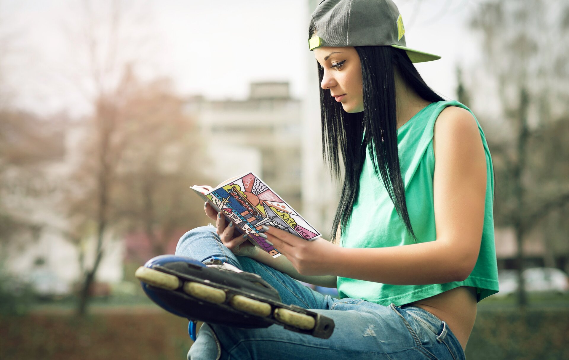 Fotografia przedstawia młodą kobietę o długich, ciemnych włosach z krótszym przodem. Kobieta jest młoda, uchwycona z profilu. Jej brwi są cienkie, a oczy podkreślone czarnym eyelinerem. Ubrana jest w krótki, zielony top bez rękawów oraz długie jeansowe spodnie. Na głowie ma szaro‑żółtą czapkę, tzw. Bejsbolówkę, z daszkiem skierowanym do tyłu. Prawą stopę podpiera o lewe kolano. Na nogach ma rolki. Siedzi na ławce, trzymając w dłoniach książkę, której okładka przedstawia kolorowy miejski krajobraz. Tło stanowi jasny budynek oraz drzewa. 