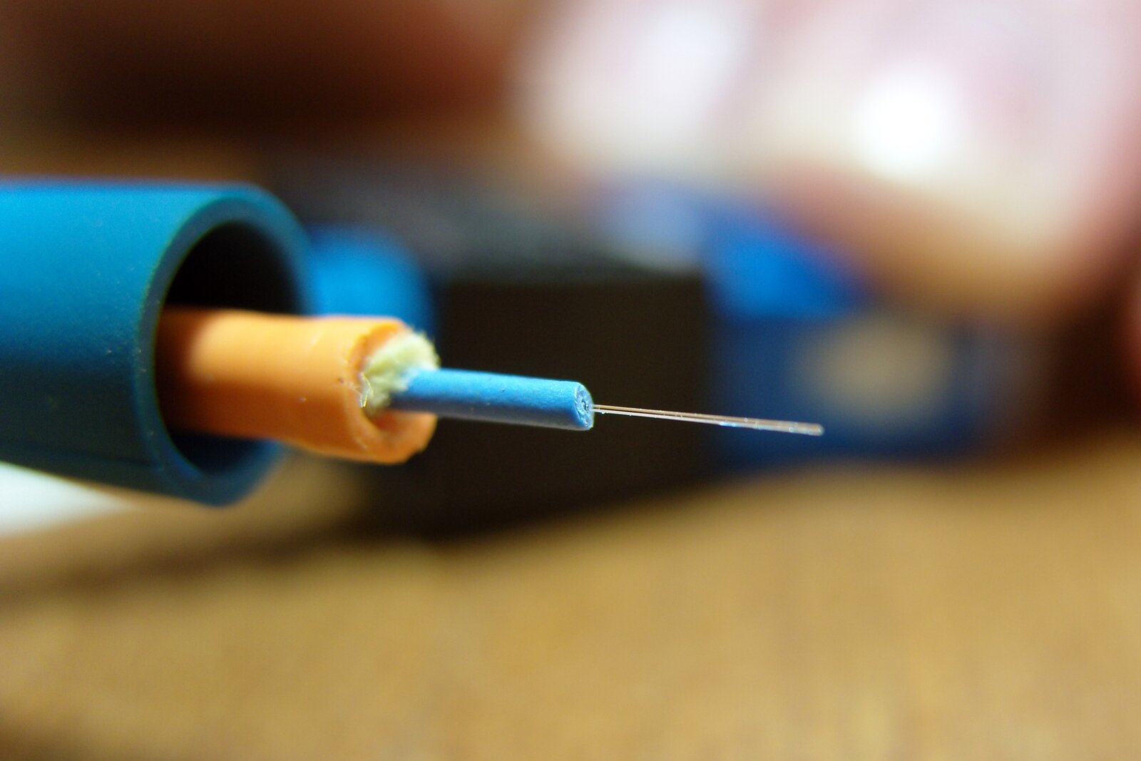 Rys. 1.  Zdjęcie przedstawia końcówkę światłowodu, z której usunięto zewnętrzne warstwy, aby ukazać jego budowę w przekroju. W dużym, niebieskim cylindrze o dość grubych ściankach umieszczono współosiowo cieńszy, żółty cylinder, a w nim jeszcze cieńszy niebieski walec. Z podstawy walca wzdłuż jego osi wychodzi cieniutki szklany pręt, który jest rdzeniem światłowodu. 