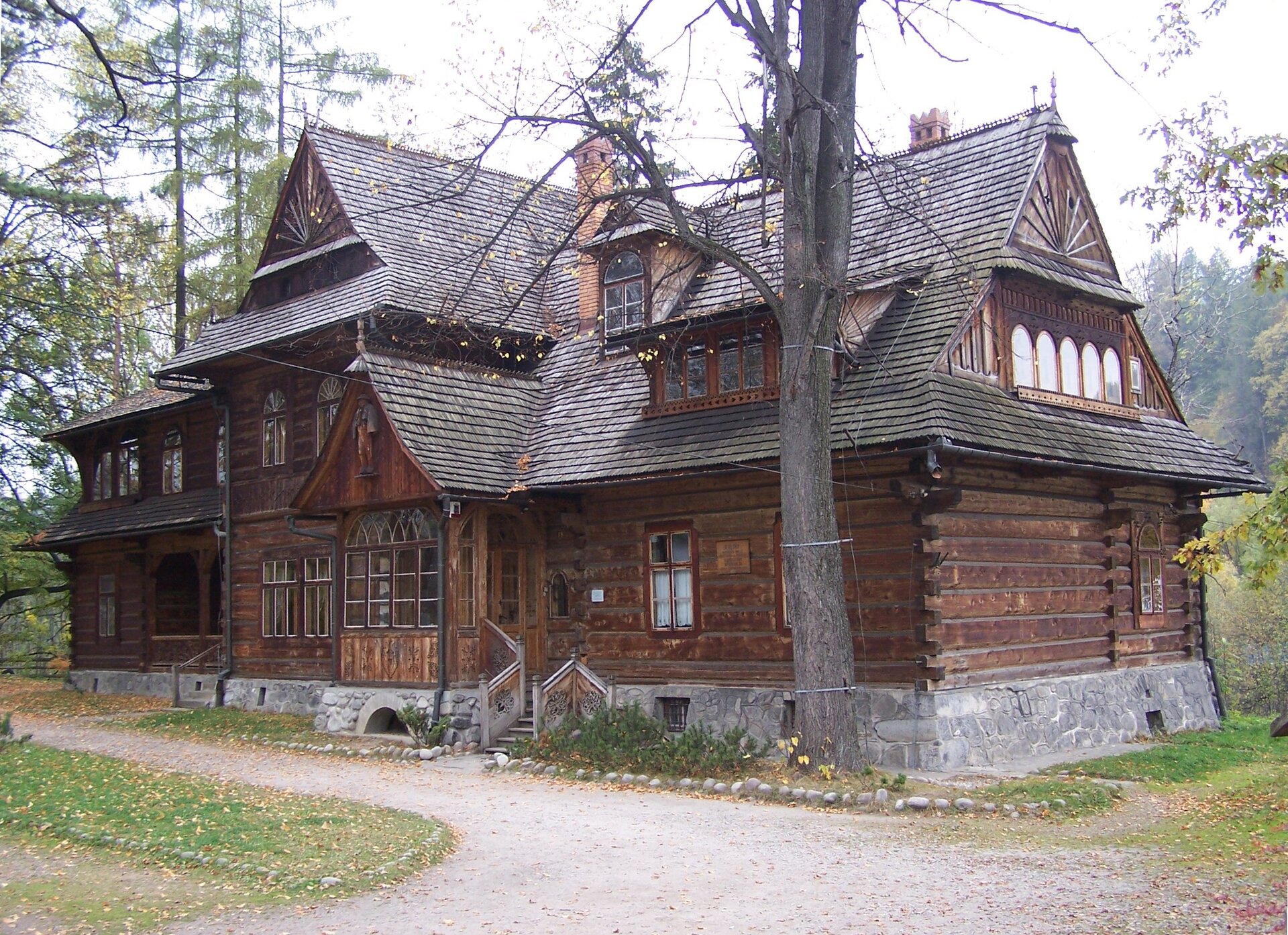 Zdjęcie przedstawia drewniany dom w góralskim stylu. Ma liczne okna - półokrągłe w górnej części. Okna są różnej wielkości. Dach jest spadzisty. Dom stoi w otoczeniu drzew.  