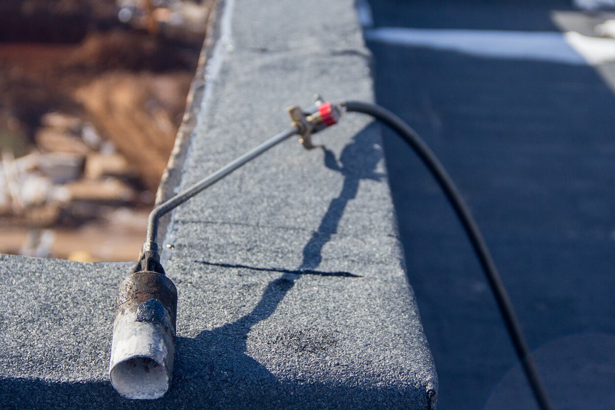 Zdjęcie przedstawia palnik do płomieniowania leżący na kamiennym murku. Narzędzie składa się z metalowego przewodu i dyszy o szerokim, okrągłym otworze, przez który kierowany jest ogień.