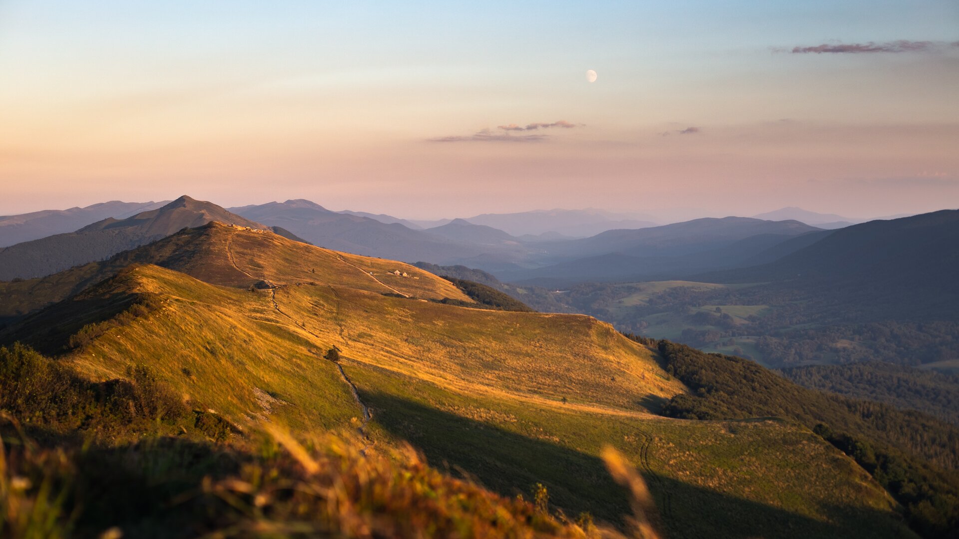 Fotografia przedstawia szczyty górskie. Na szczycie znajdują się polany, poniżej nich drzewa. W oddali widać kolejne pasma górskie.
