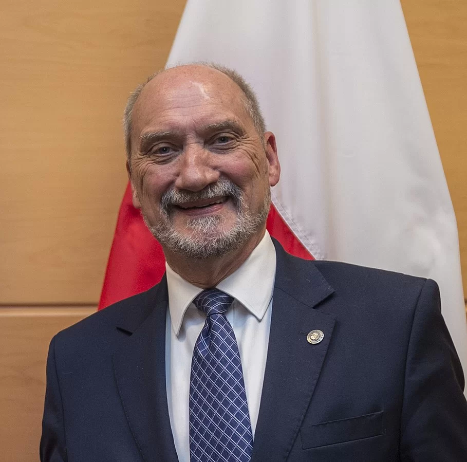 Na zdjęciu znajduje się Antoni Macierewicz. Jest to starszy mężczyzna z krótką, siwą brodą i o krótkich włosach. Jest ubrany w ciemny garnitur. Mężczyzna ma zmarszczki na twarzy, uśmiecha się. Za nim widoczna jest flaga Polski.