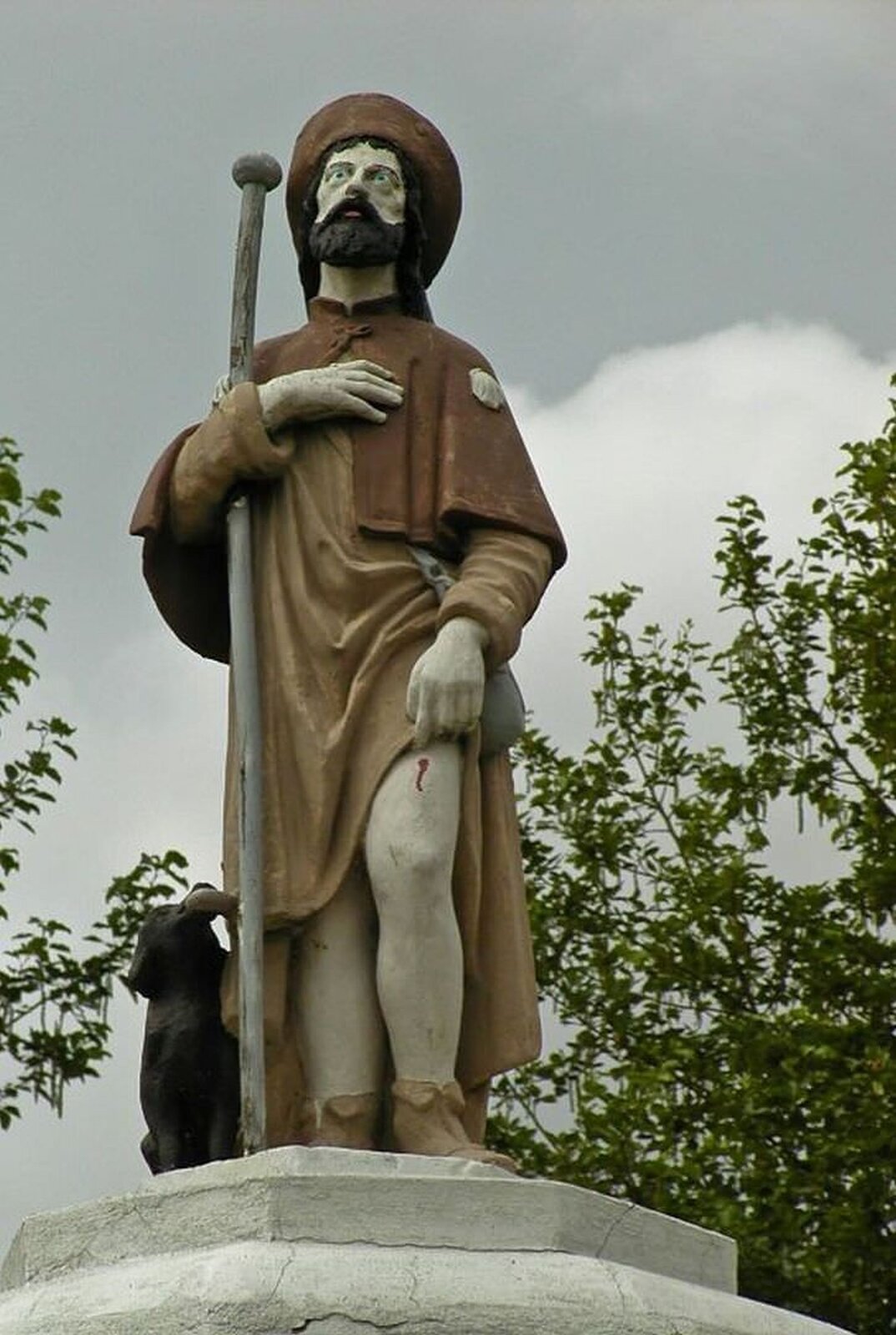 lustracja przedstawia figurę świętego Rocha. Święty w dłoni trzyma laskę, a obok niego siedzi pies. Na głowie ma brązowe nakrycie i szatę w takim samym kolorze.