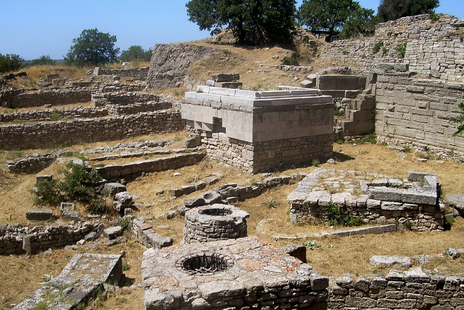 Zdjęcie przedstawia kamienne konstrukcje, pomiędzy którymi rośnie wyschnięta trawa. Kamienie są płaskie i dobrze do siebie dopasowane, w niektórych miejscach widać wygładzane prostokątne kamienie, które łączone były za pomocą jakiejś zaprawy o jednolitym kolorze. Widoczne są dwie koliste konstrukcje, najprawdopodobniej studnie, z czego w jednej wbudowanej w większy prostokątny blok, widać kratę wykonaną z metalu.
