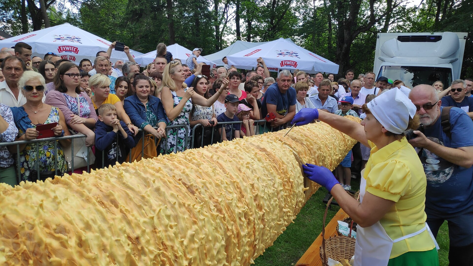 Zdjęcie przedstawia kobietę krojącą olbrzymiego sękacza i tłum ludzi przyglądający się jej. Kobieta ubrana jest w żółtą koszulkę na krótki rękaw  oraz zieloną spódnicę. Ma na sobie ochronny biały fartuch, białą chustę na głowie oraz na dłoniach w których trzyma nóż, ma niebieskie rękawiczki jednorazowe. Kobietę po prawej stronie fotografuje starszy mężczyzna  w czarnych okularach z siwą brodą. Tłum ludzi stojący naprzeciwko ubrany jest w lekką wiosenną odzież, próbuje  uwiecznić chwile  z wydarzenia przy użyciu telefonów komórkowych. W tle można dostrzec  białe parasole oraz białe auto dostawcze.