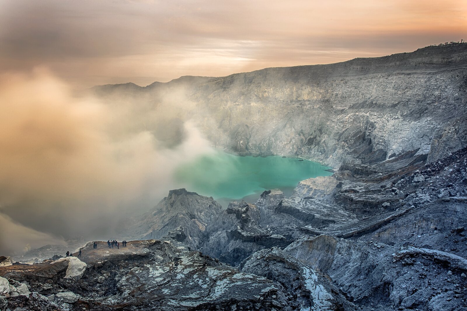 Zdjęcie przedstawia kopalnię siarki Kawah Ijen w Indonezji. Krater wulkanu, w którym znajduje się woda jezioro częściowo zasłania gęsty dym będący oparami gazów wydobywających się z jeszcze aktywnego wulkanu.