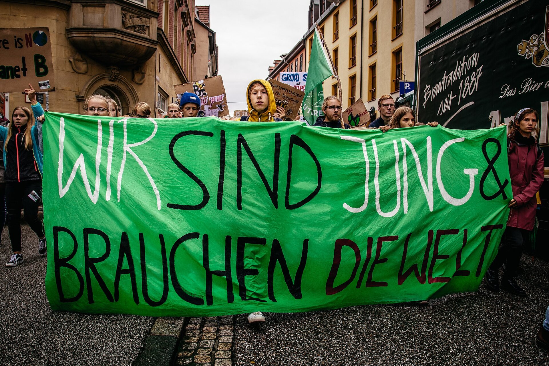 Zdjęcie przedstawia protest w obronie klimatu. Młodzi ludzie na pierwszym planie trzymają przed sobą transparent z napisem Wir sind jung und brauchen die Welt.