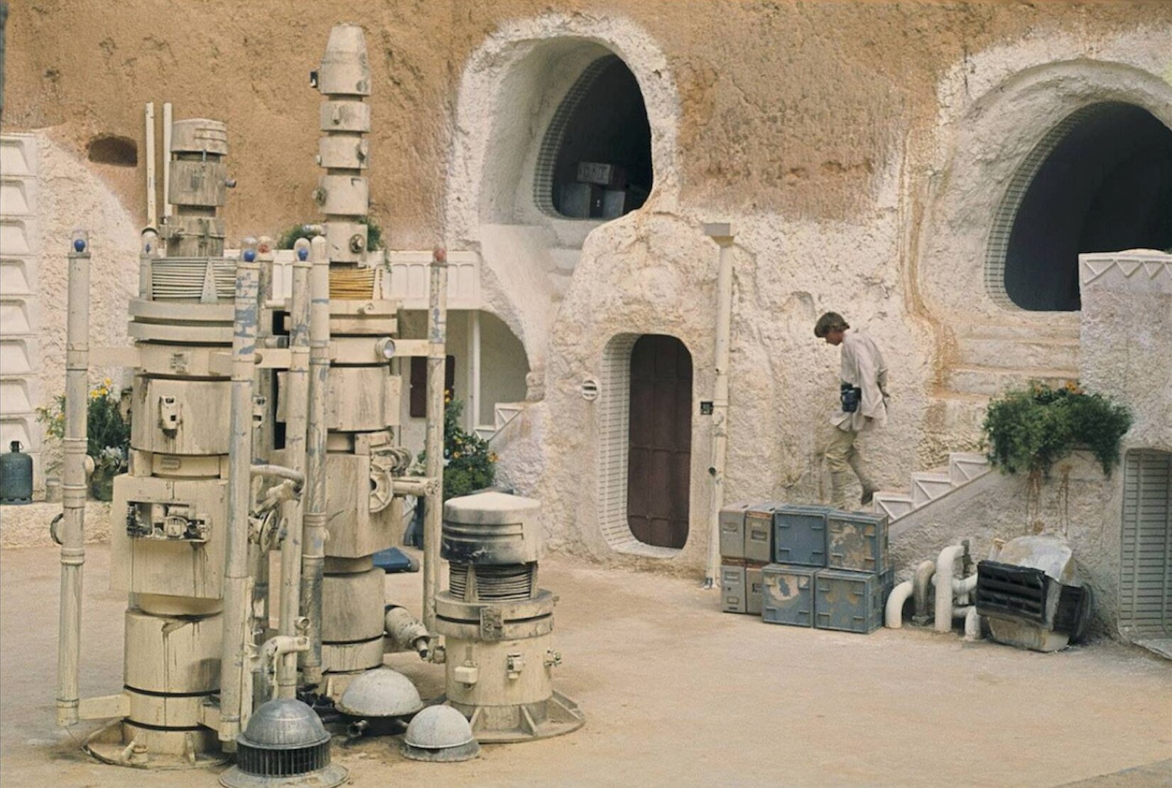 Ilustracja przedstawia Hotel Sidi Driss w Matmacie w Tunezji. Jest to rzeczywista lokalizacja akcji filmu „Gwiezdne Wojny” z 1977 roku. Na pierwszym planie, z lewej strony znajdują się 3 roboty w kolorze kremowym. 2 z nich są wysokie, a jeden niski. Na drugim planie, z prawej strony ilustracji znajduje się mężczyzna schodzący po schodach. Ma on blond włosy i ubrany jest w kremową bluzkę z długim rękawem i z czarnym pasem w talii, kremowe spodnie oraz kremowe buty. Przy schodach stoją niebieskie skrzynki, obok nich znajdują się kremowe rury i średniej wielkości przedmiot w kształcie koła. Jest on koloru piaskowego, a na środku znajdują się czarne elementy. Nad nim jest zielona roślinność. W tle ilustracji jest ściana budynku hotelowego w kolorze piaskowym. Na górze wykute są 2 otwory, a na dole znajdują się brązowe drzwi. Po lewej stronie ilustracji, za robotami, znajduje się zielona roślinność z żółtymi kwiatami oraz średniej wielkości zielona butla. 