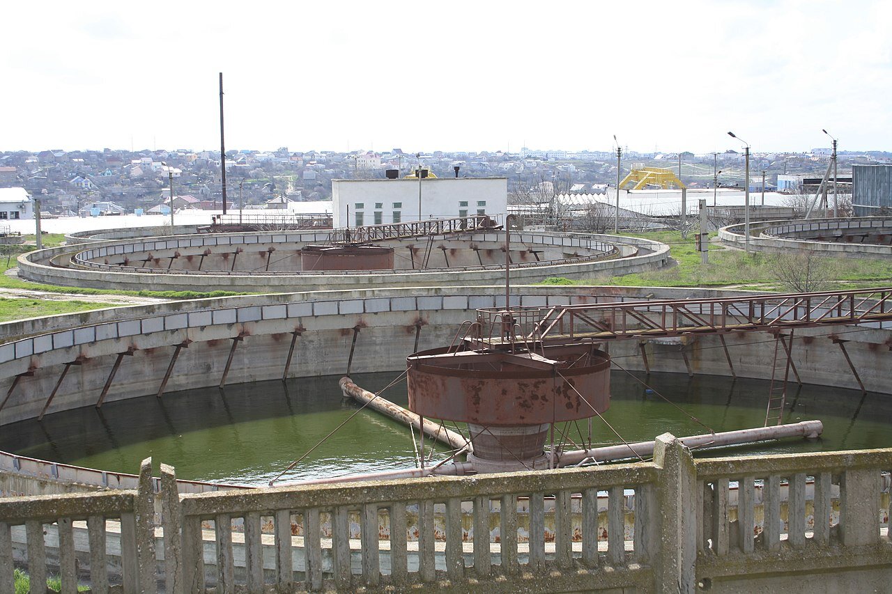 Zdjęcie przedstawia osadniki oczyszczalni ścieków. Konstrukcje mają kształt ogromnych, betonowych okręgów wypełnionych wodą. Na środku każdej konstrukcji znajduje się metalowy walec z czterema ramionami.