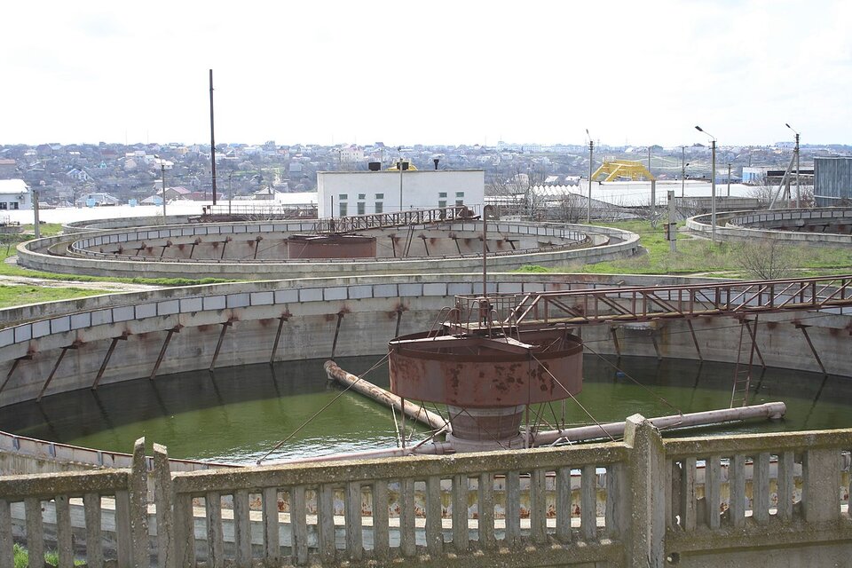Zdjęcie przedstawia osadniki oczyszczalni ścieków. Konstrukcje mają kształt ogromnych, betonowych okręgów wypełnionych wodą. Na środku każdej konstrukcji znajduje się metalowy walec z czterema ramionami.