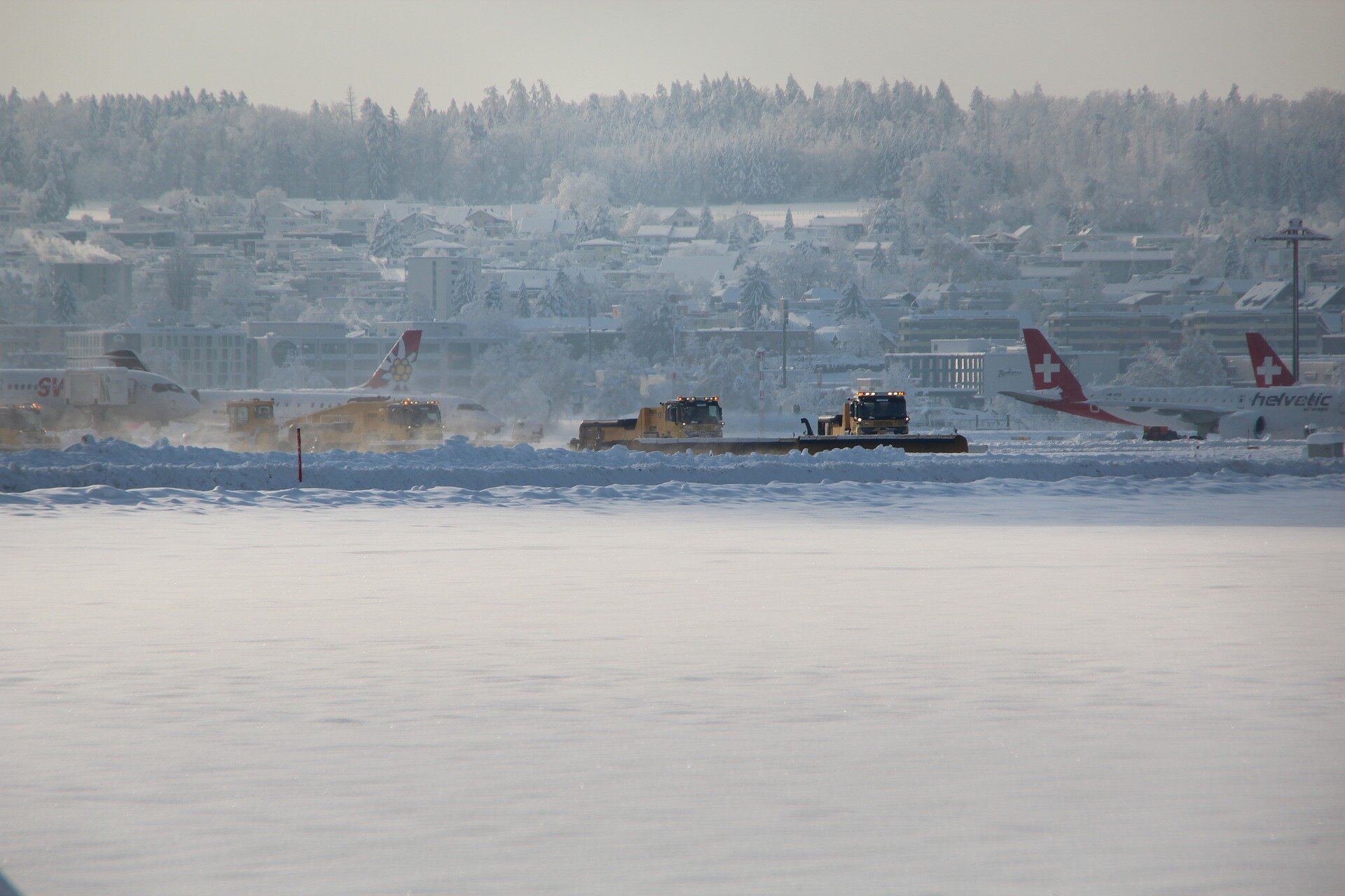 Zdjęcie przedstawiające kilka ciągników siodłowych wraz z doczepionymi z przodu pługami śnieżnymi usuwającymi grubą warstwę śniegu z płyty lotniska. Na drugim planie samoloty oraz budynki.