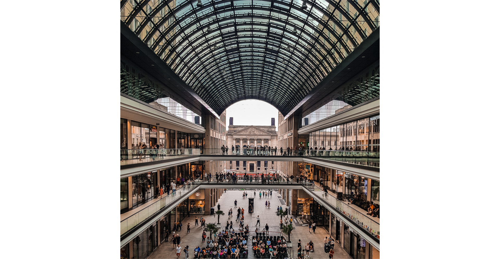Zdjęcie zostało zrobione wewnątrz centrum handlowego. Widać zatłoczony parter ze sklepami po obu stronach holu. Pierwsze i drugie piętro również ma sklepy po obu stronach budynku, klienci przemieszczają się po tarasach umieszczonych wokół otwartej przestrzeni na wskroś całego budynku. Widoczny jest półokrągły dach centrum handlowego. W oddali rozpościera się otwarty dziedziniec i budynek stojący za centrum.