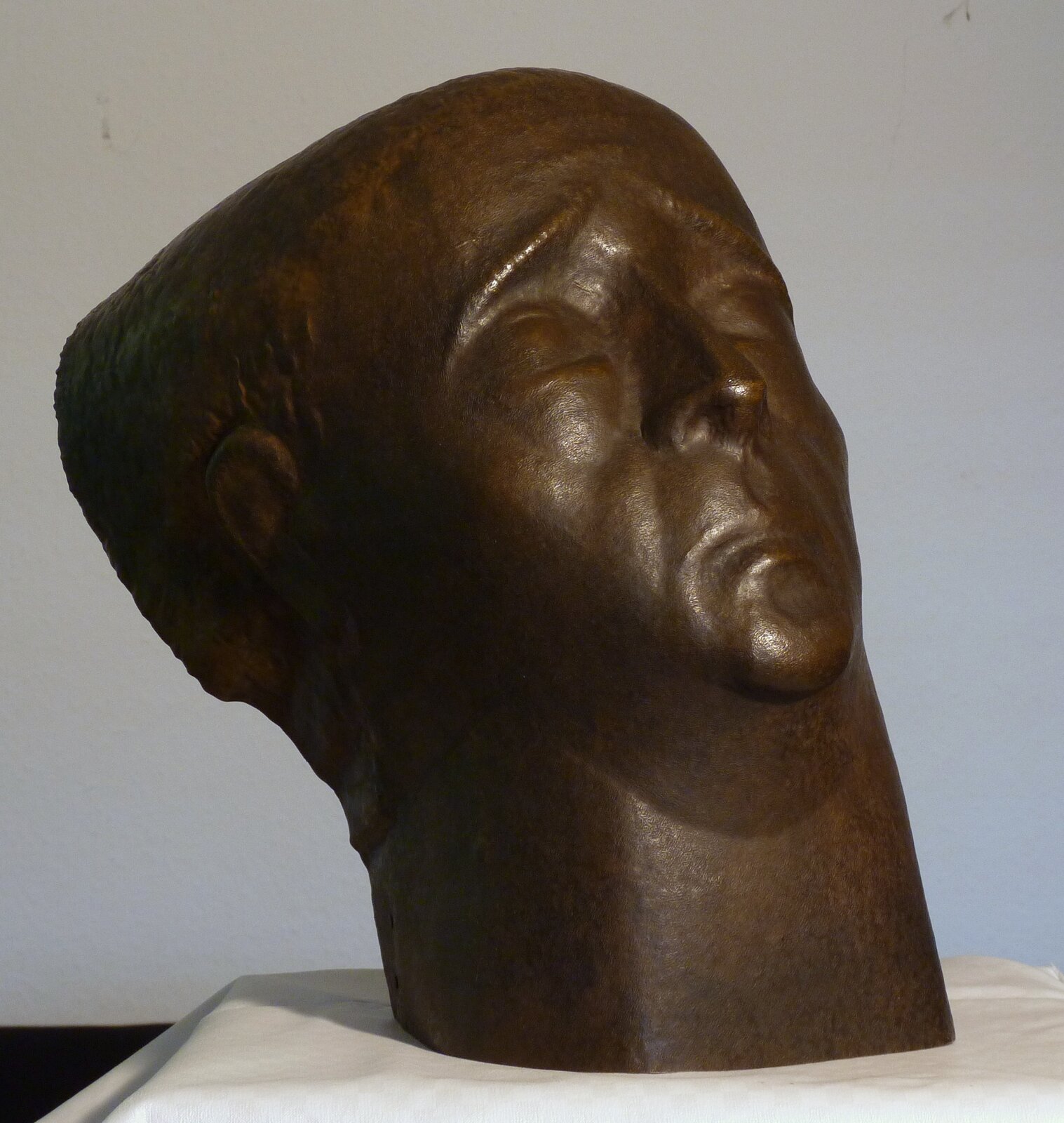 Zdjęcie przedstawia rzeźbę. Jest to wykonana z mosiądzu ludzka głowa z szeroko otwartymi oczami, wysoko postawionymi brwiami i zmarszczonym czołem. Spojrzenie jest skierowane w górę. Tył głowy jest nienaturalnie wystający. W tle jest biała ściana.