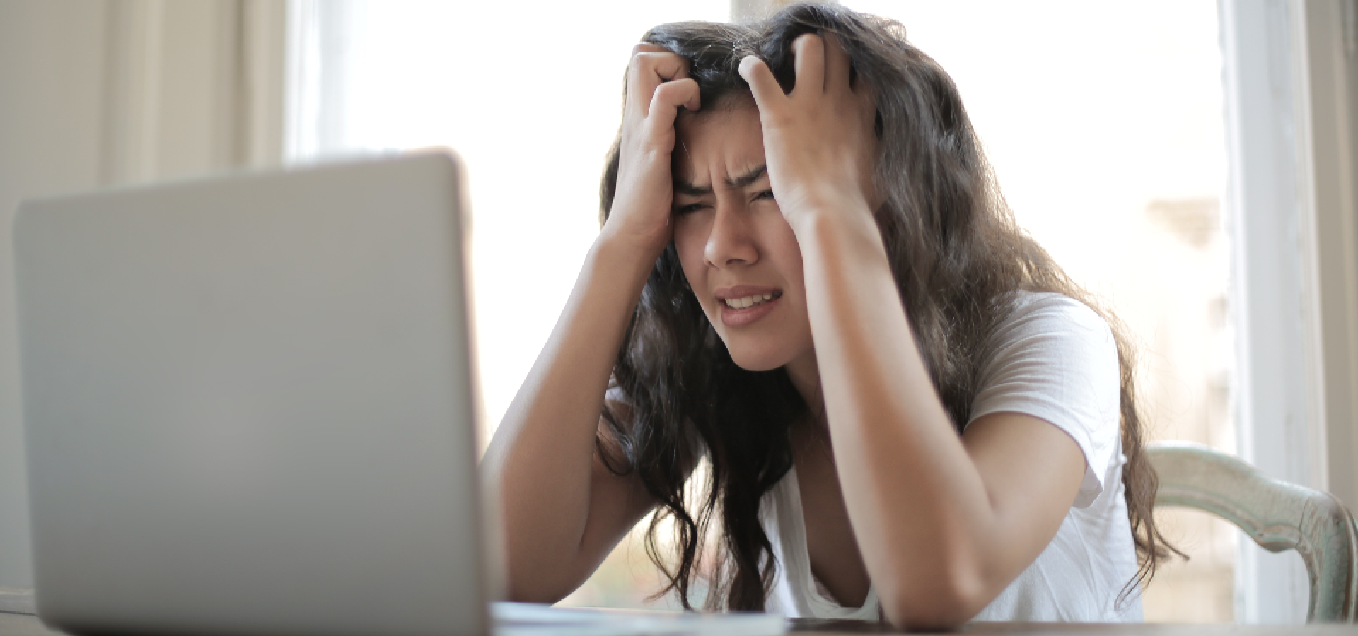 Zdjęcie przedstawia dziewczynę, która siedzi przy komputerze. Trzyma się za głowę dwoma rękami i ma poirytowaną minę. 