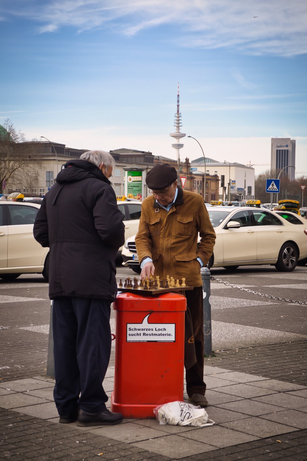 Zdjęcie przedstawia dwóch starszych mężczyzn grających w szachy. Mężczyźni znajdują się na chodniku, a szachy rozstawione są na metalowym słupku, na którym widnieje napis Schwarze Loch sucht Restmaterie. Obok mężczyzn przejeżdżają ulicą samochody osobowe. W tle znajdują się budynki i wieża.