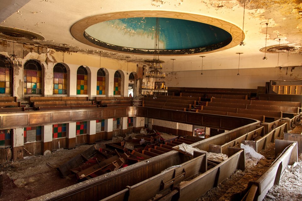 Na zdjęciu jest wnętrze teatru. W suficie znajduje się duża okrągła wnęka, z której zwisa ozdobny żyrandol. Na poziomie balkonów stoją rzędy ławek. Poniżej, na poziomie podstawowym także stoją ławki, część z nich jest poprzewracana. Panuje duży bałagan, leżą duże ilości odrapanej farby.