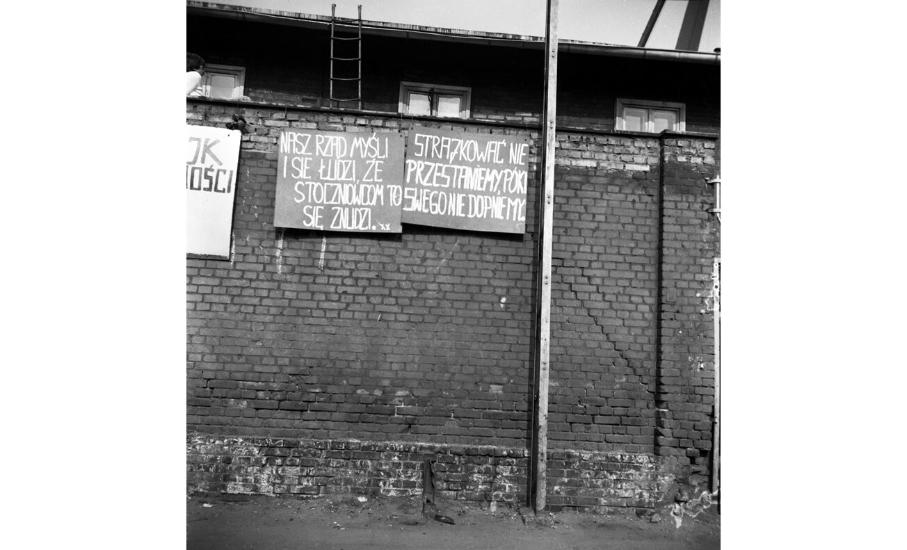 Czarno-biała fotografia przedstawiająca transparenty wywieszone na murze stoczni w Gdańsku. Znajdują się na nich hasła "strajkować nie przestaniemy, póki swego nie dopniemy".