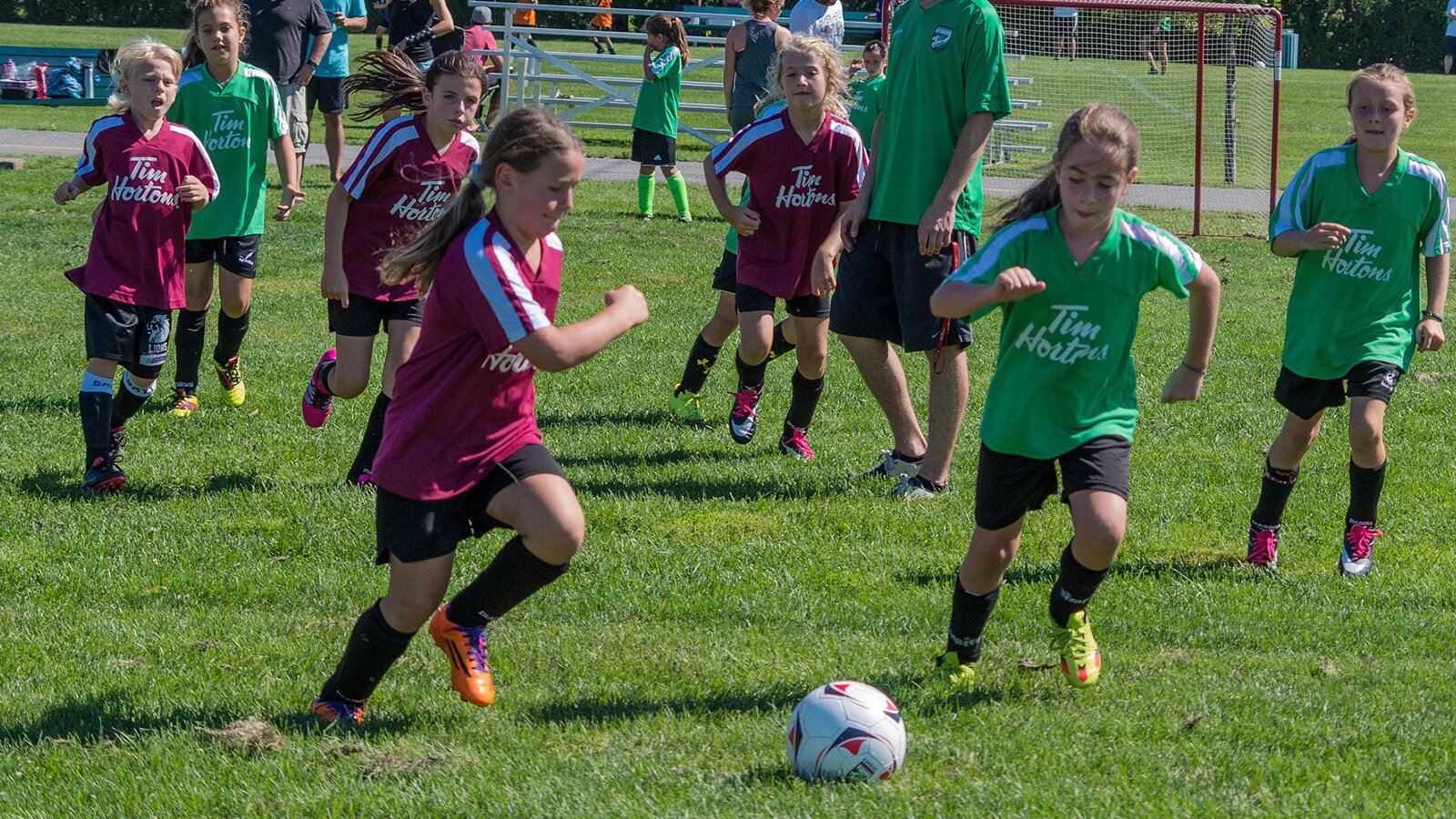 Zdjęcie przedstawia dwie drużyny dzieci w wieku około 11 lat grające w piłkę nożną. Dzieci są ubrane  w stroje piłkarskie. Jedna drużyna ma zielone koszulki i czarne szorty, a druga czerwone koszulki i czarne szorty. Większość grupy to dziewczynki. Na pierwszym planie widoczne są dwie dziewczynki biegnące za piłką.