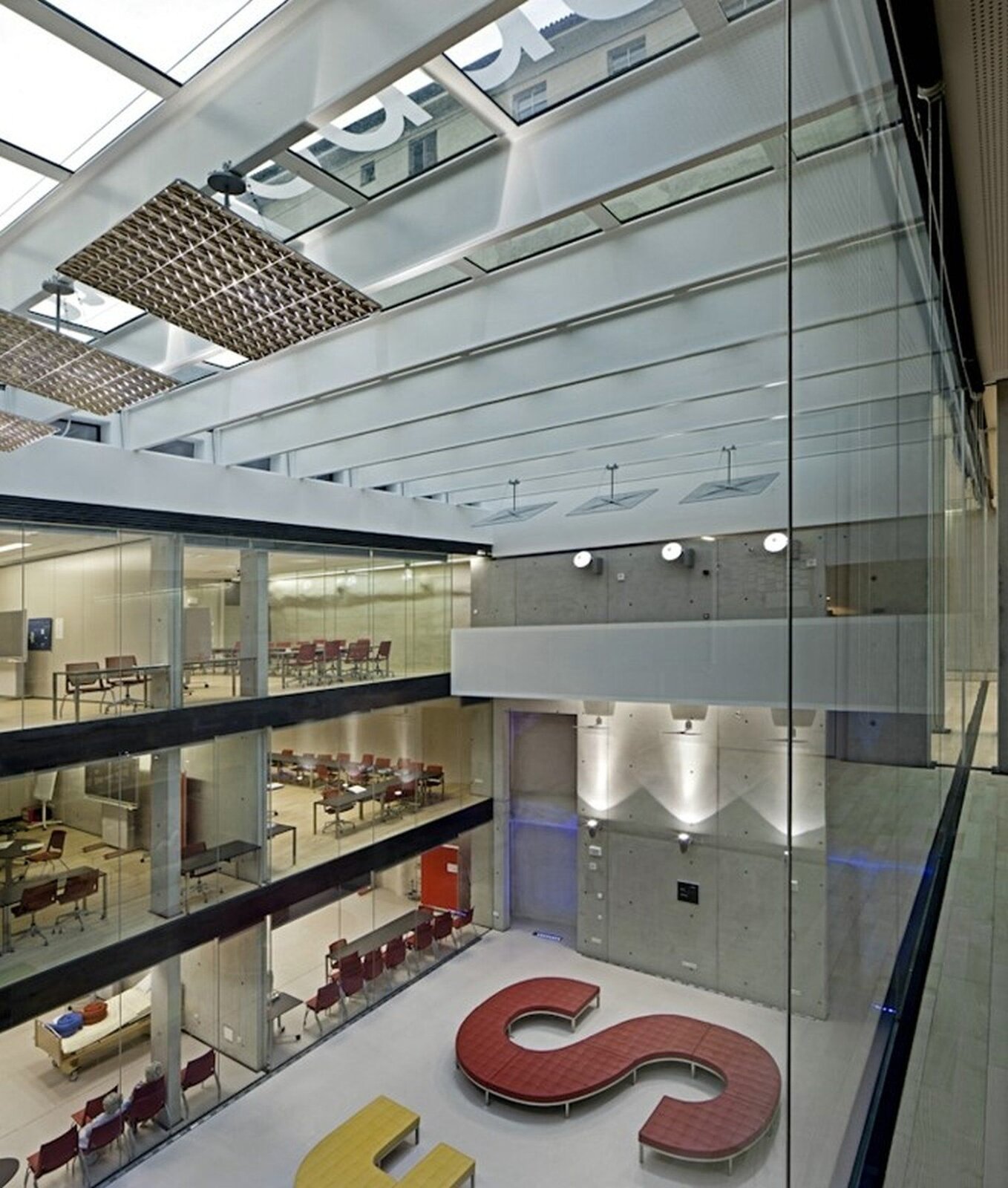 Ilustracja przedstawia widok na podziemną szkołę w Bolzano. Na zdjęciu ukazane są trzy kondygnacje. Ściany są wykonane ze szkła. W każdej z widocznych sal znajdują się krzesła. Przed pomieszczeniami znajduje się przestronny hol ze szklanym dachem. Z prawej strony ulokowana jest winda ze stalowymi drzwiami.