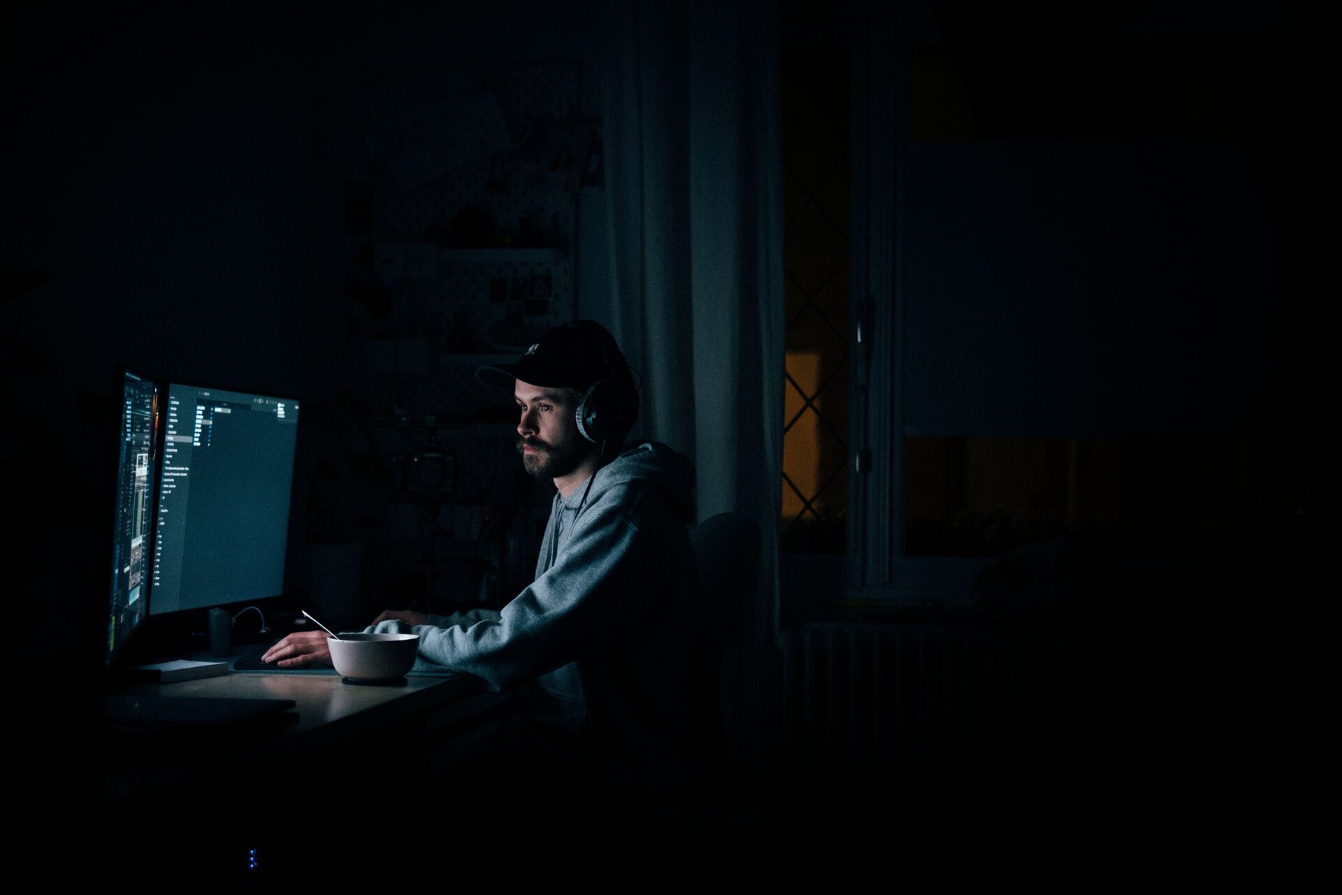 Zdjęcie przedstawia mężczyznę siedzącego na krześle przy biurku. Jedynym źródłem światła jest poświata monitora. Mężczyzna ma na sobie czapkę z daszkiem i słuchawki. Na biurku widoczna też jest miseczka z łyżką. Transkrypcja nagrań znajduje się poniżej
