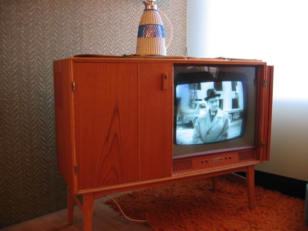Zdjęcie przedstawia starodawny telewizor. Jego wypukły szklany ekran znajduje się w drewnianej szafce. Pod nim jest pomarańczowy dywan. W tle jest wzorzysta, brązowa tapeta.