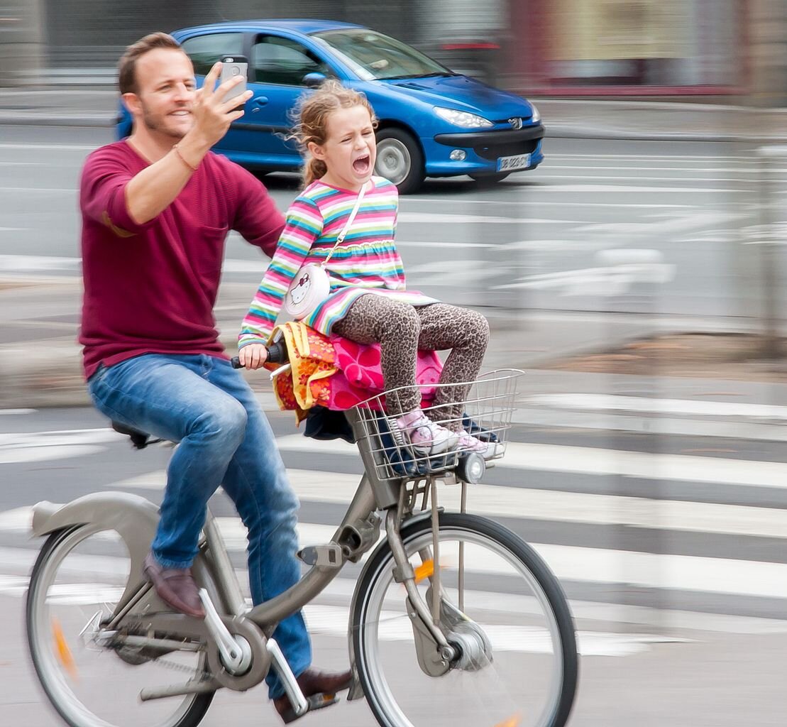 Zdjęcie przedstawia mężczyznę jadącego na rowerze. Przed nim, na kierownicy roweru, z nogami na koszyku przymocowanym nad przednim kołem pojazdu siedzi dziewczynka. Jej oczy są zamknięte, natomiast usta szeroko rozwarte. Mężczyzna uśmiech się. Jego jedna ręka jest uniesiona, a w dłoni znajduje się telefon. 