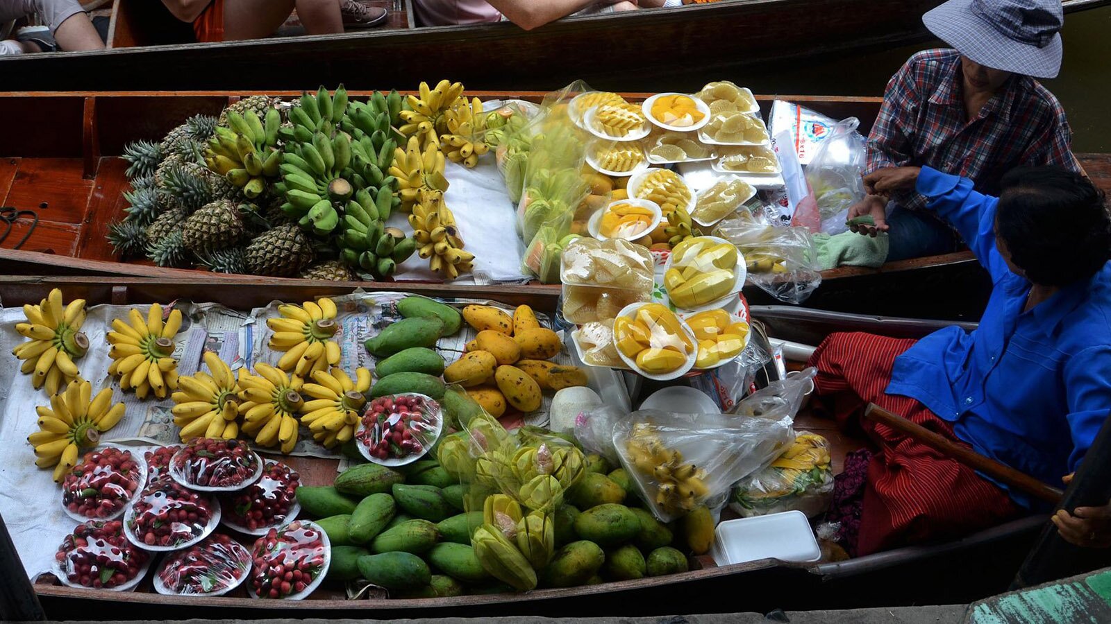 Zdjęcie przedstawia dwie łodzie płynące obok siebie, w każdej siedzi jedna osoba. Lodzie wypełnione są owocami, widoczne są: ananasy, banany w kiściach żółte i zielone, papaja, mango. Te same owoce obrane i pokrojone ułożono na talerzykach i zakryto folią spożywczą.